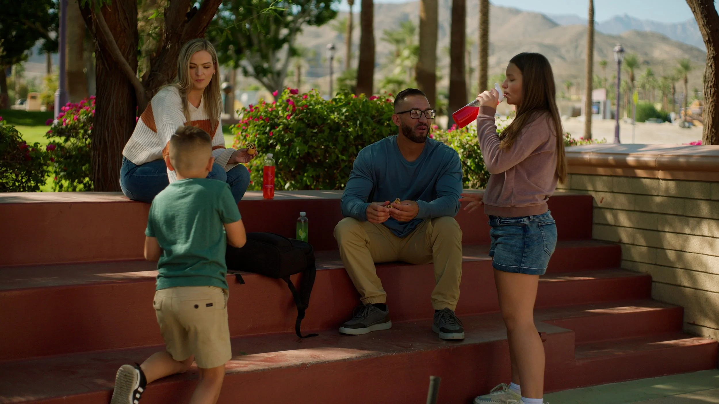 Group of four people relaxing on outdoor steps in a park setting, with two adults sitting and two children standing, one drinking from a cup.