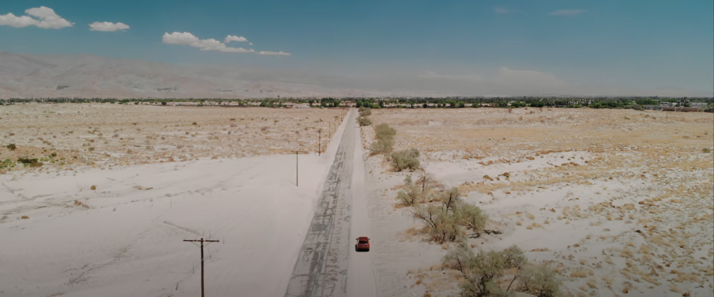 Desert landscape with a long road, a red car driving, and distant mountains.