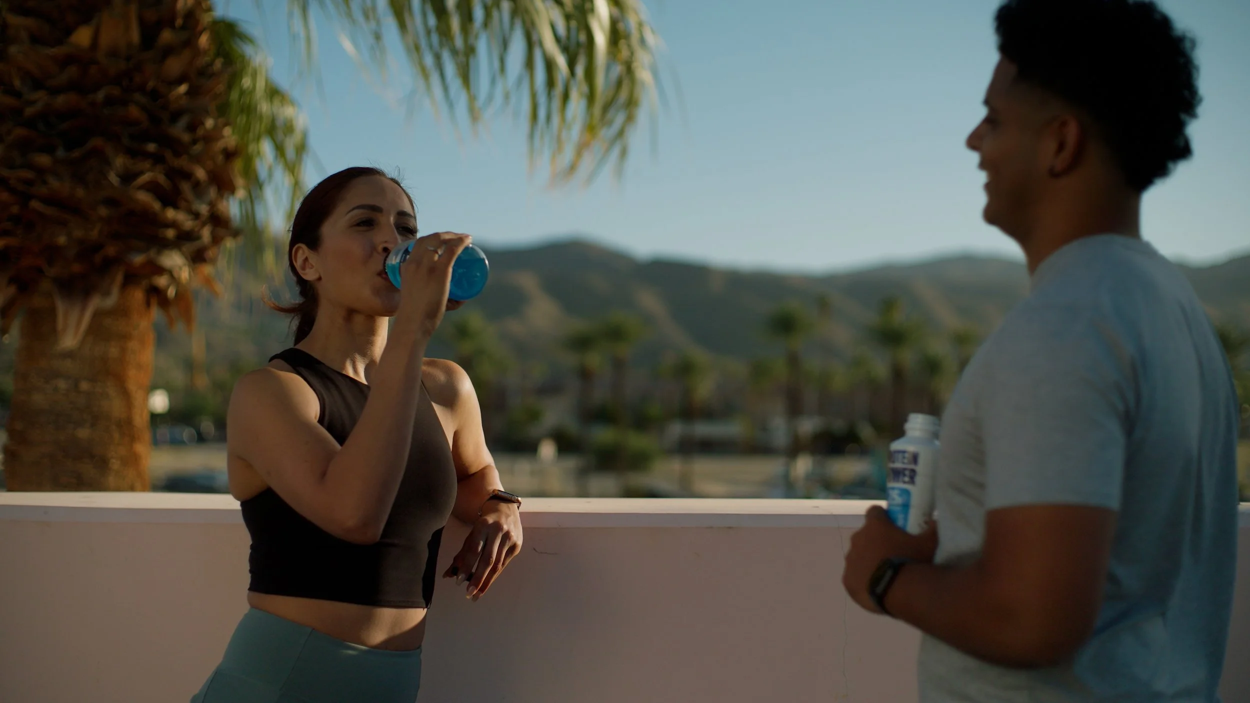 Two people outdoors drinking fitness beverages, one with a blue drink and the other with a protein shake, near a palm tree with mountains in the background.