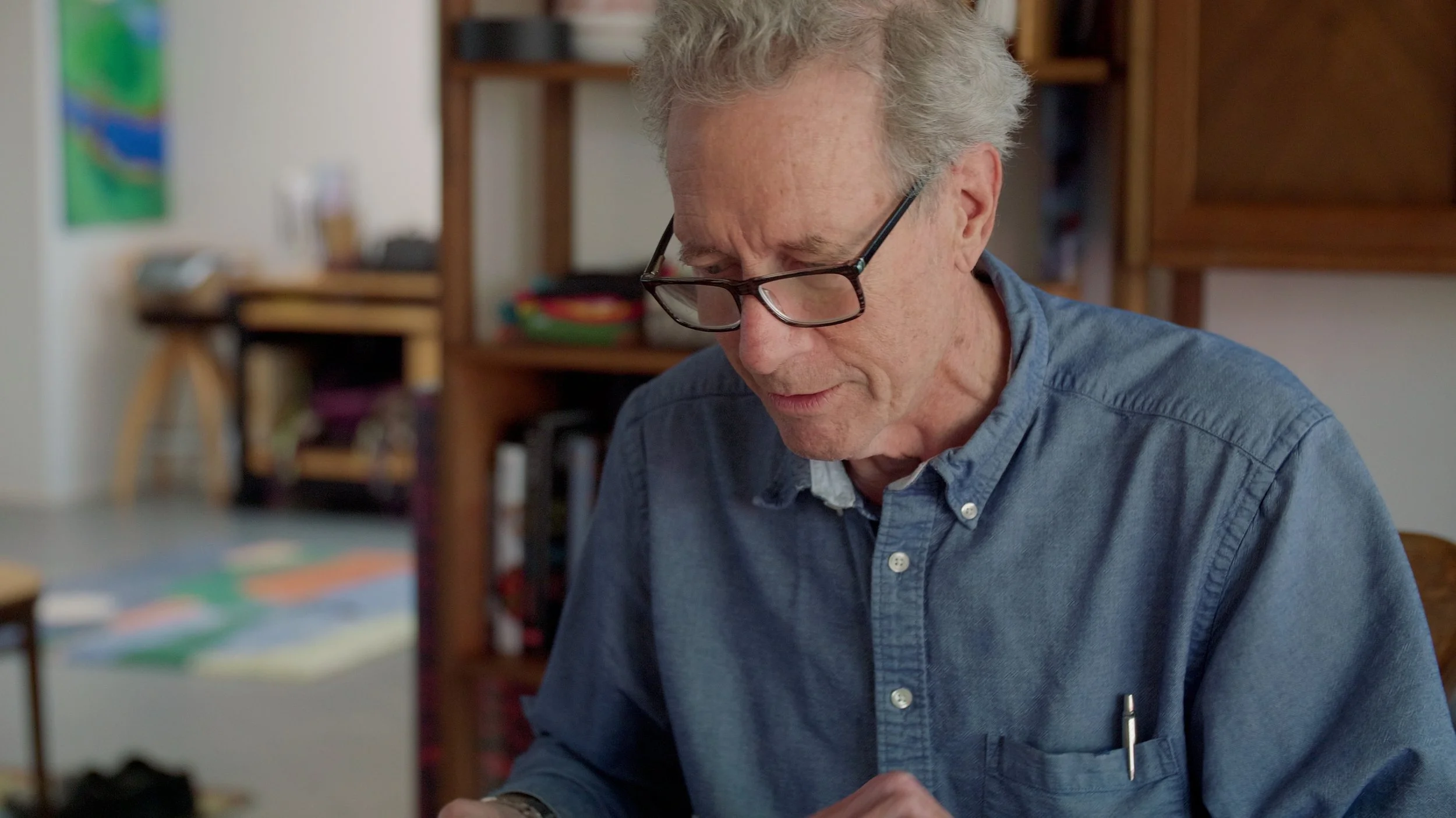 Man wearing glasses and a blue shirt, sitting indoors, focused and reading, with bookshelves and colorful décor in the background.