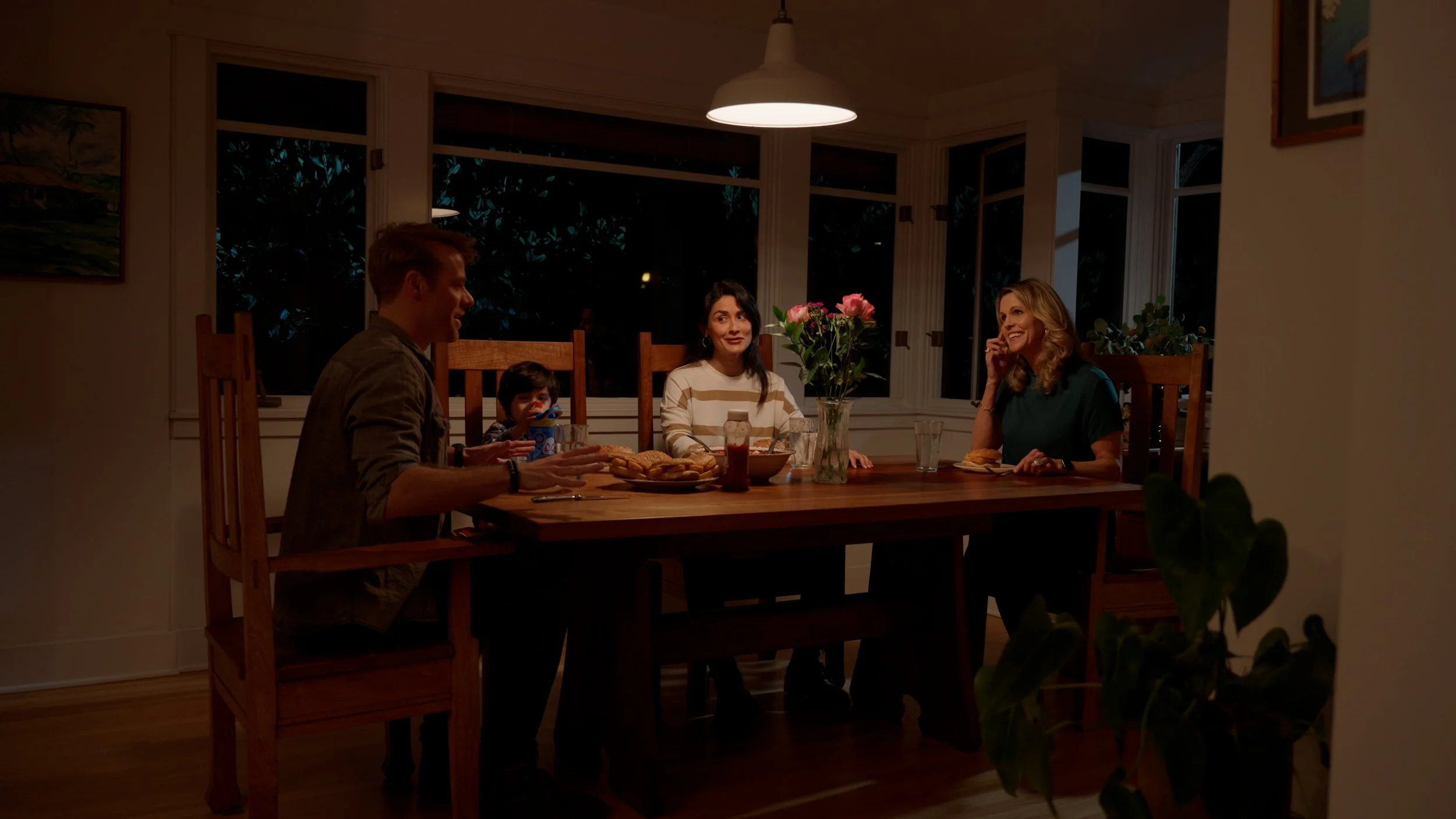 Four people sitting at a dining table sharing a meal, surrounded by windows, in a dimly lit room. A vase with pink flowers is on the table.