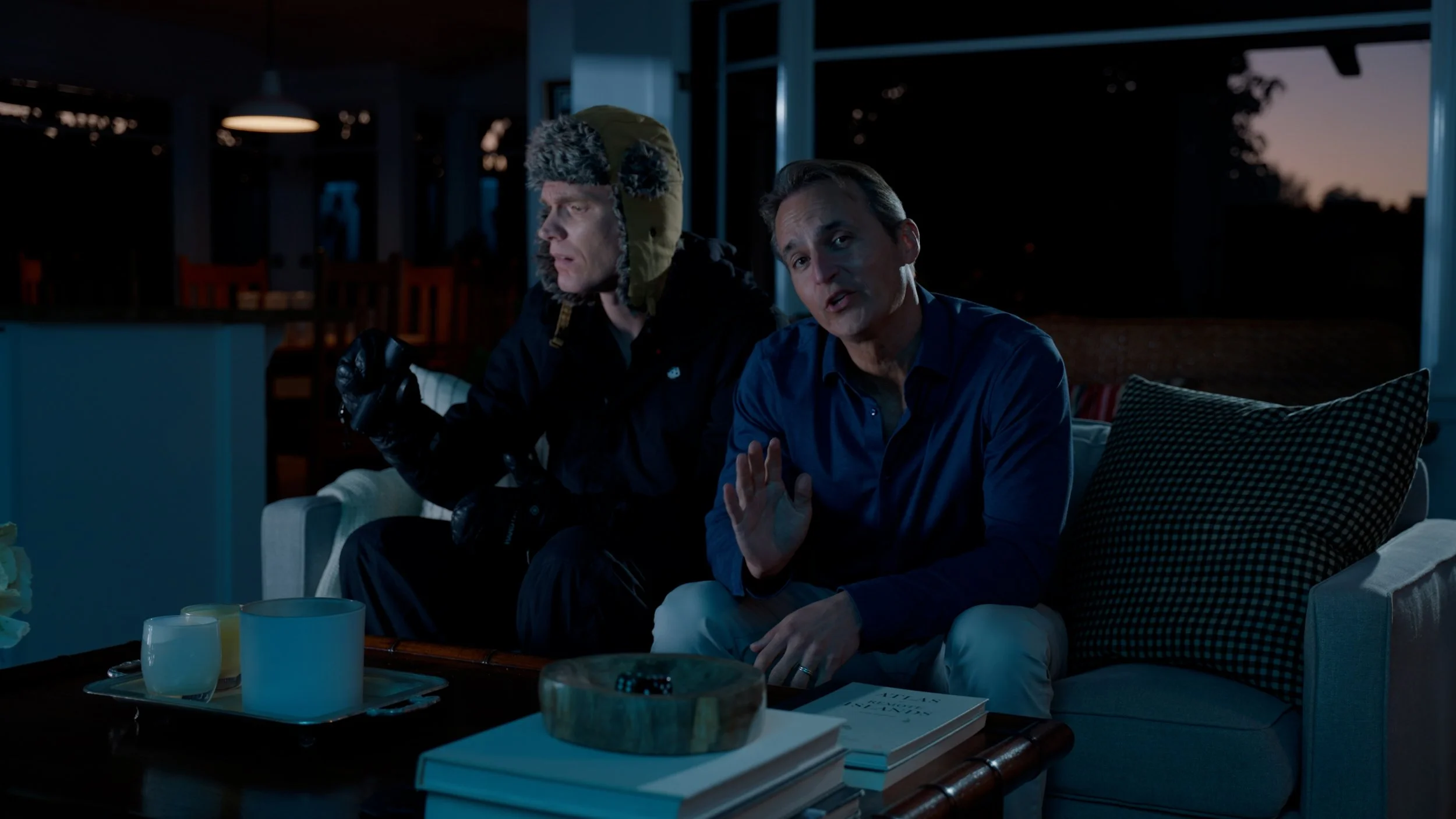 Two men sitting on a couch at night, one wearing a winter hat and gloves, the other in a blue shirt, with books and drinks on a table nearby.