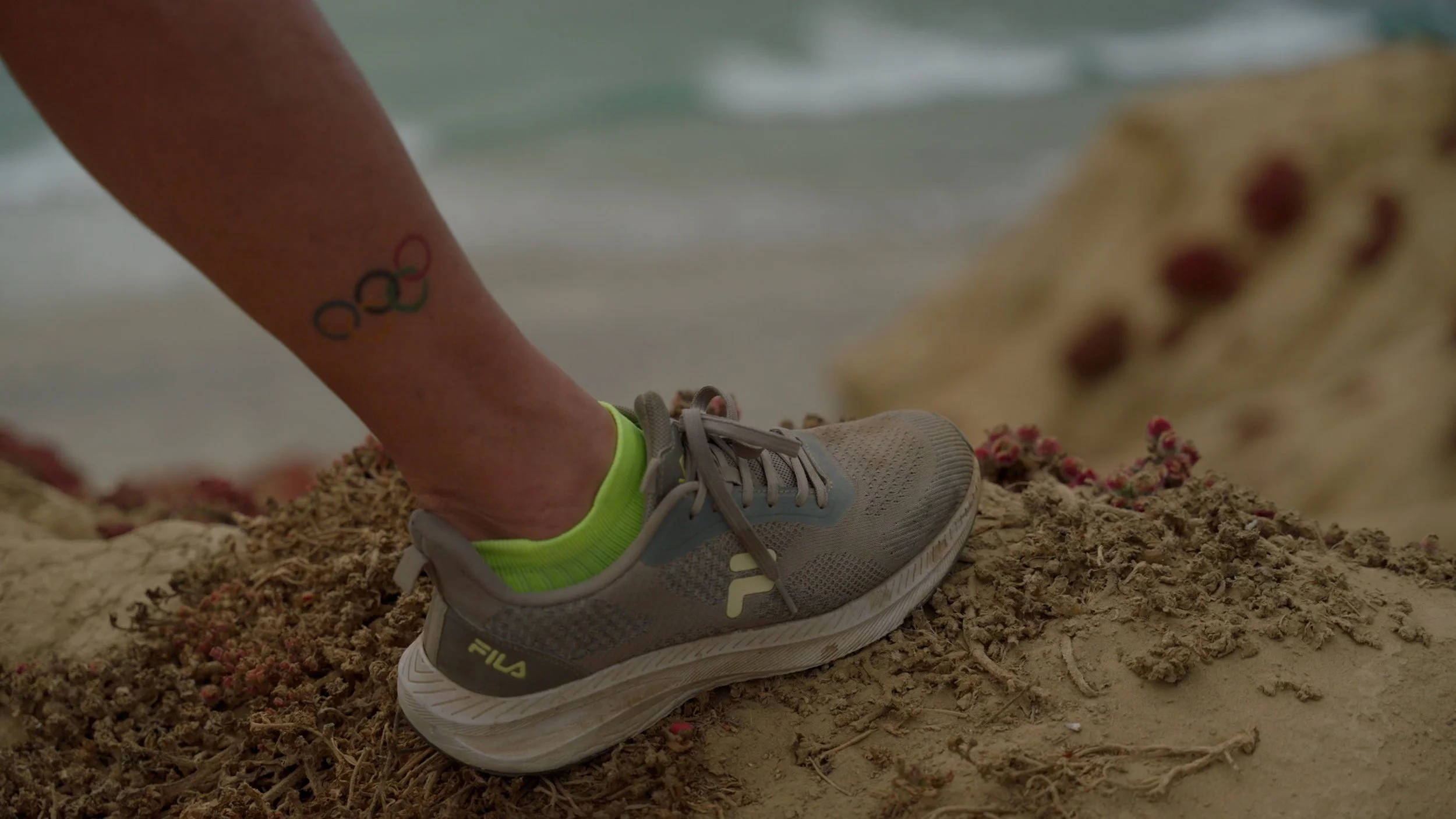 Person wearing Fila shoe on sandy terrain with Olympic rings tattoo on ankle.