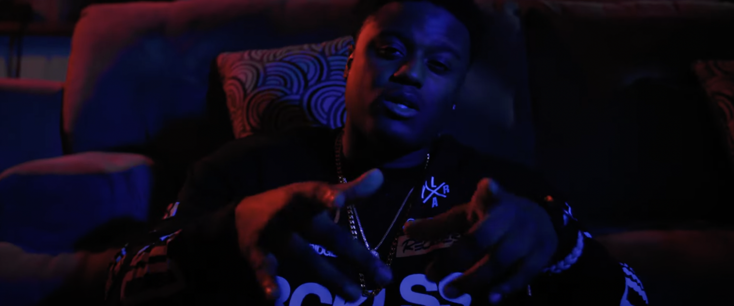 Person sitting on a couch in dim lighting, wearing a black shirt with white text and a gold chain, gesturing with hands.