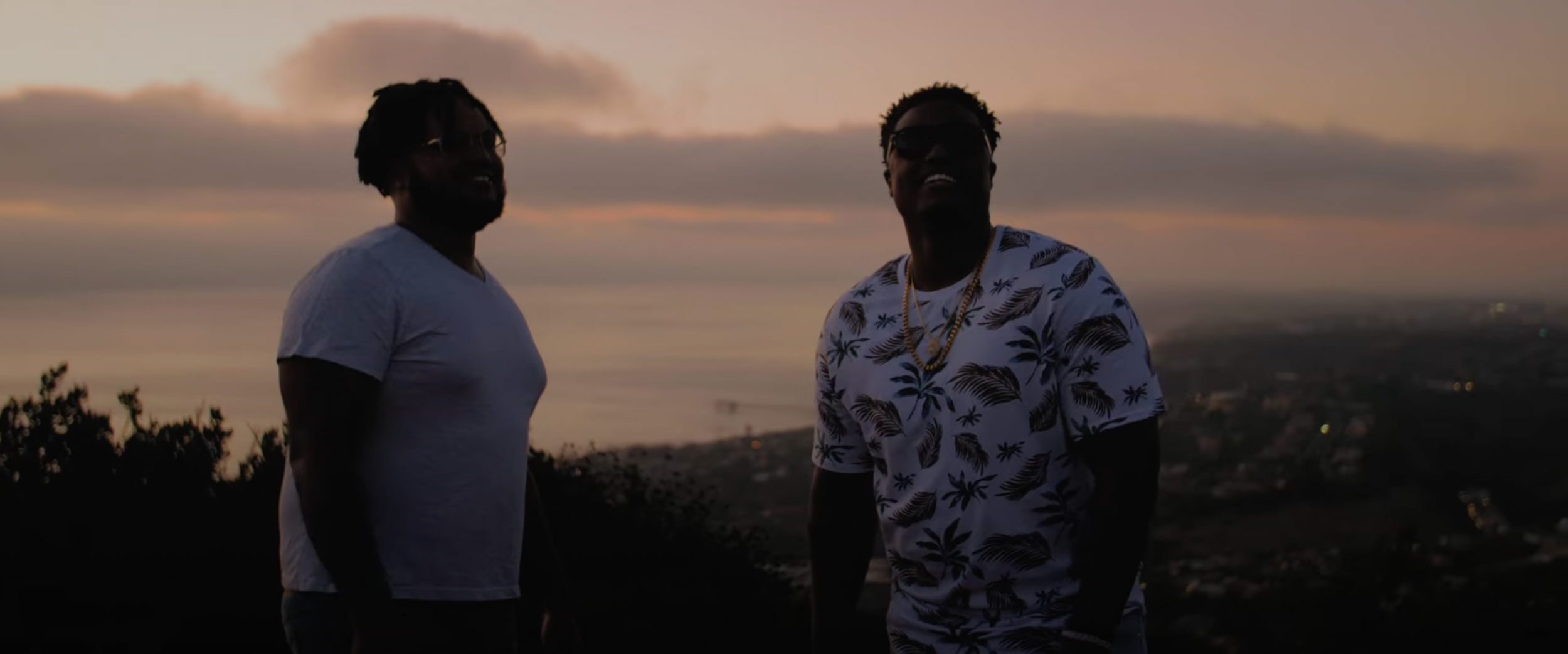 Two men standing in front of a scenic sunset with ocean view, wearing casual summer outfits.