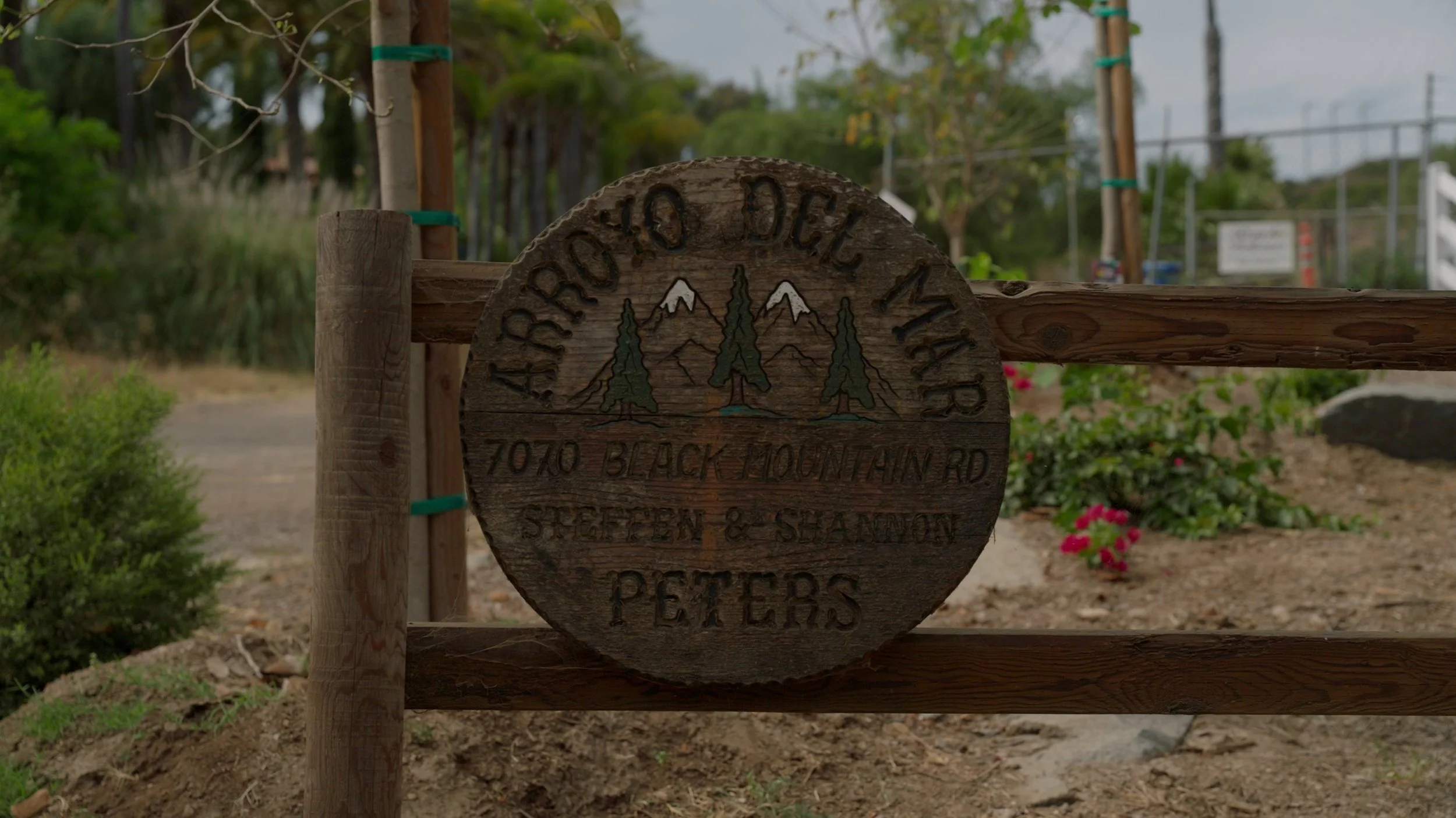 Wooden sign reading 'Arroyo Del Mar' with address '7070 Black Mountain Rd' and names 'Steffen & Shannon Peters'; decorative trees and mountains.