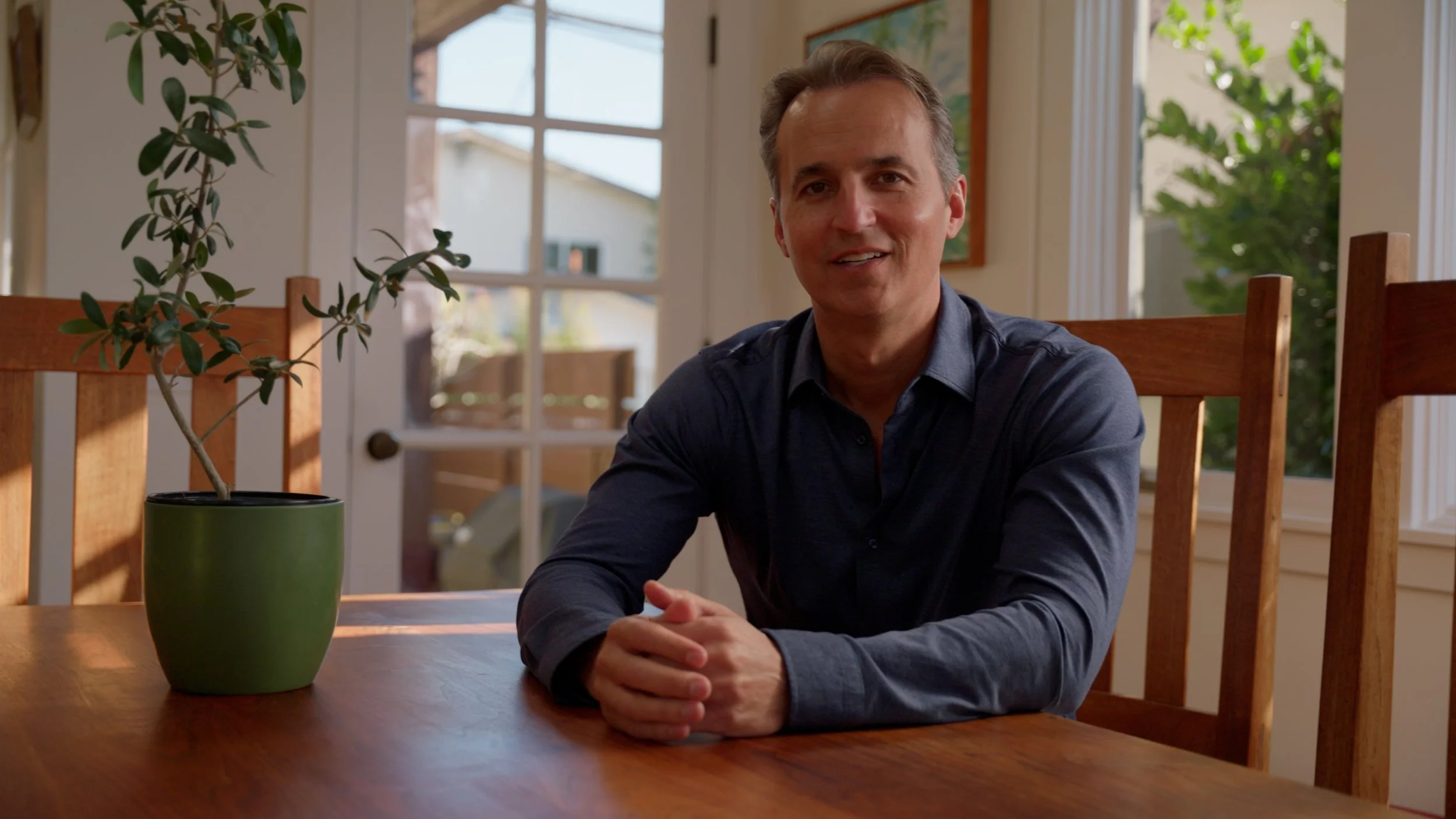 Person sitting at a wooden table with a potted plant, indoors, near a window.