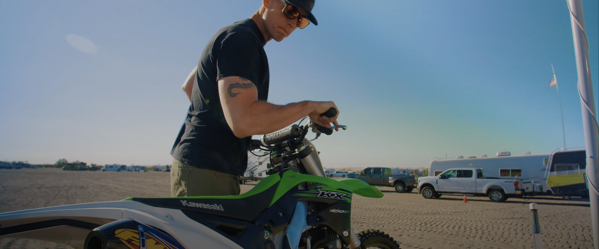 Man with tattoo adjusting Kawasaki dirt bike at campsite