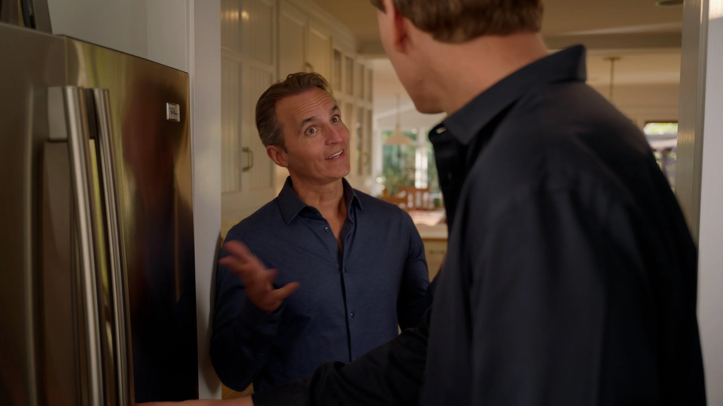 Two men conversing in a kitchen next to a stainless steel refrigerator.