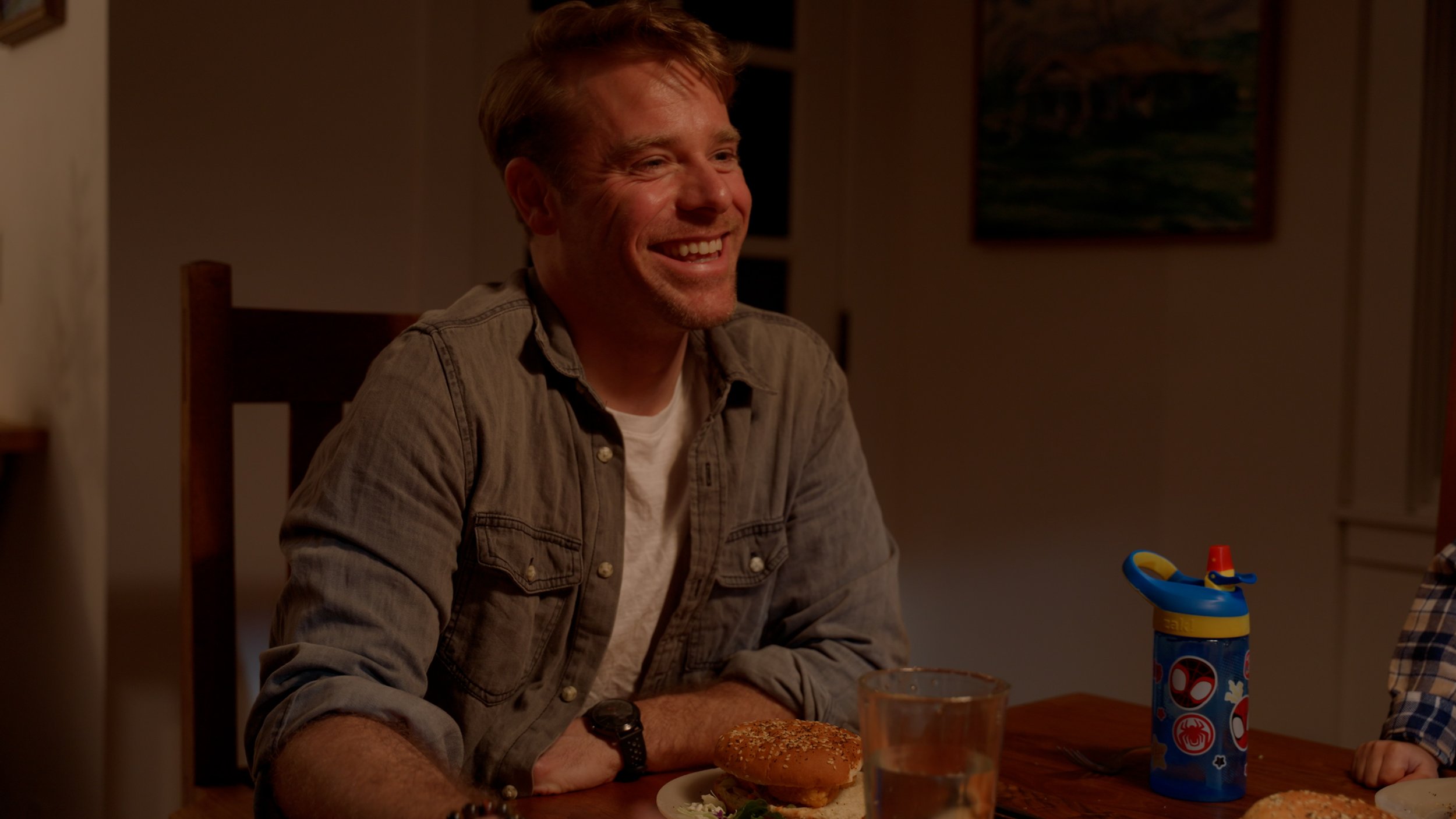 Smiling man sitting at a table with a burger and a child's water bottle.