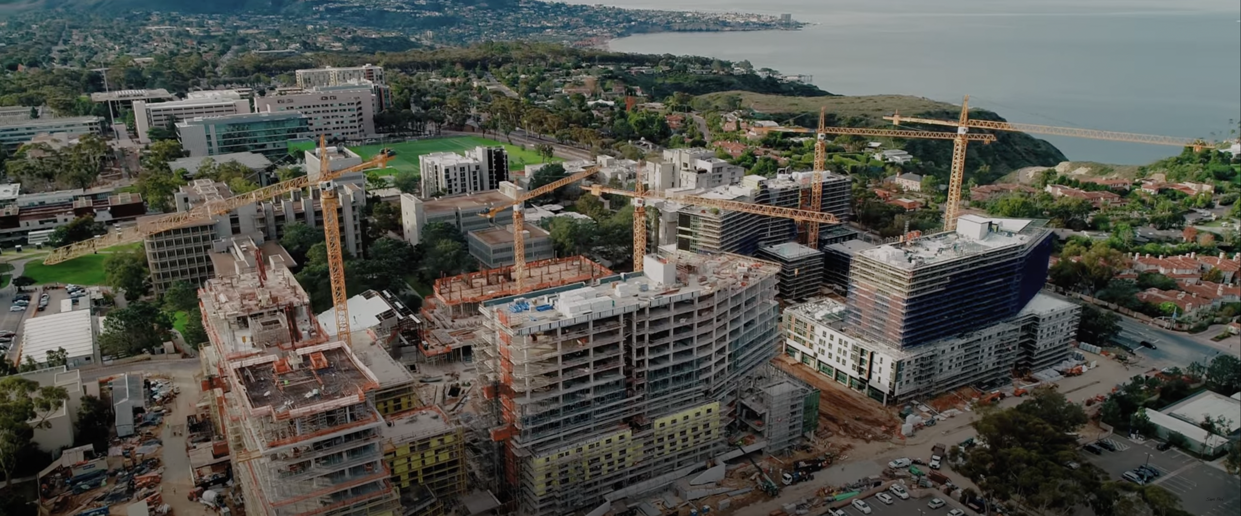 Aerial view of a construction site with multiple cranes and partially constructed buildings, surrounded by a green landscape and a body of water in the background.