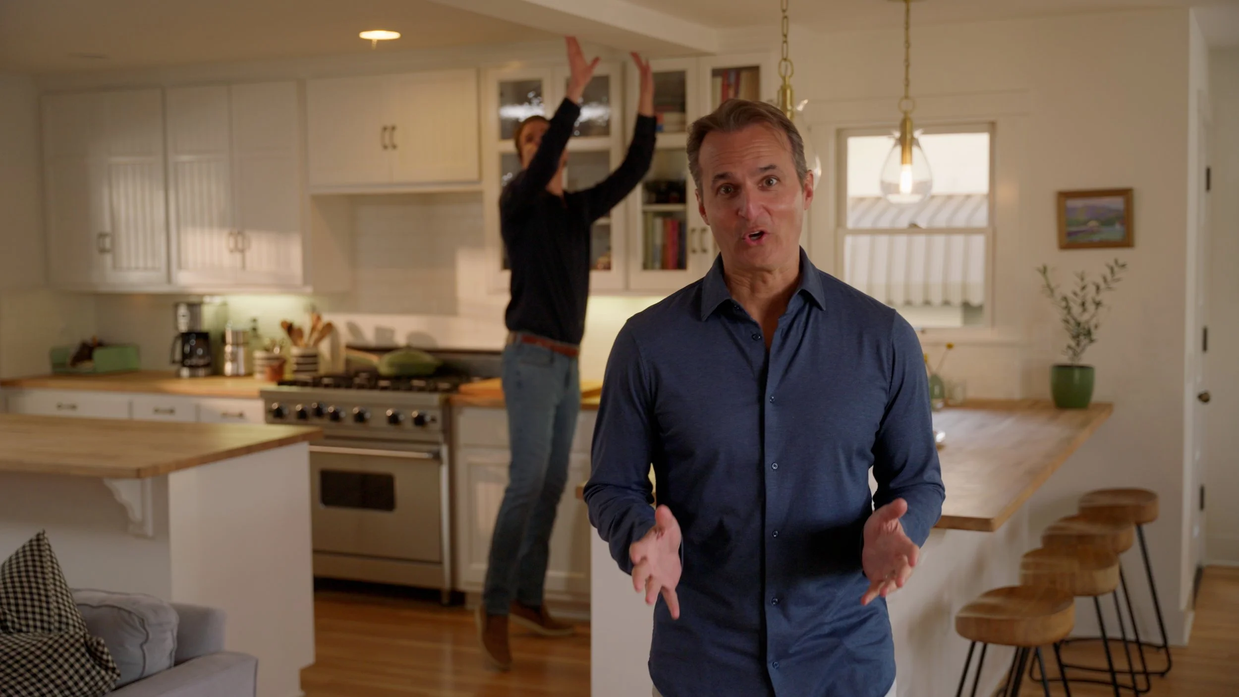 Two people in a modern kitchen; one speaking in the foreground, the other reaching for a shelf.