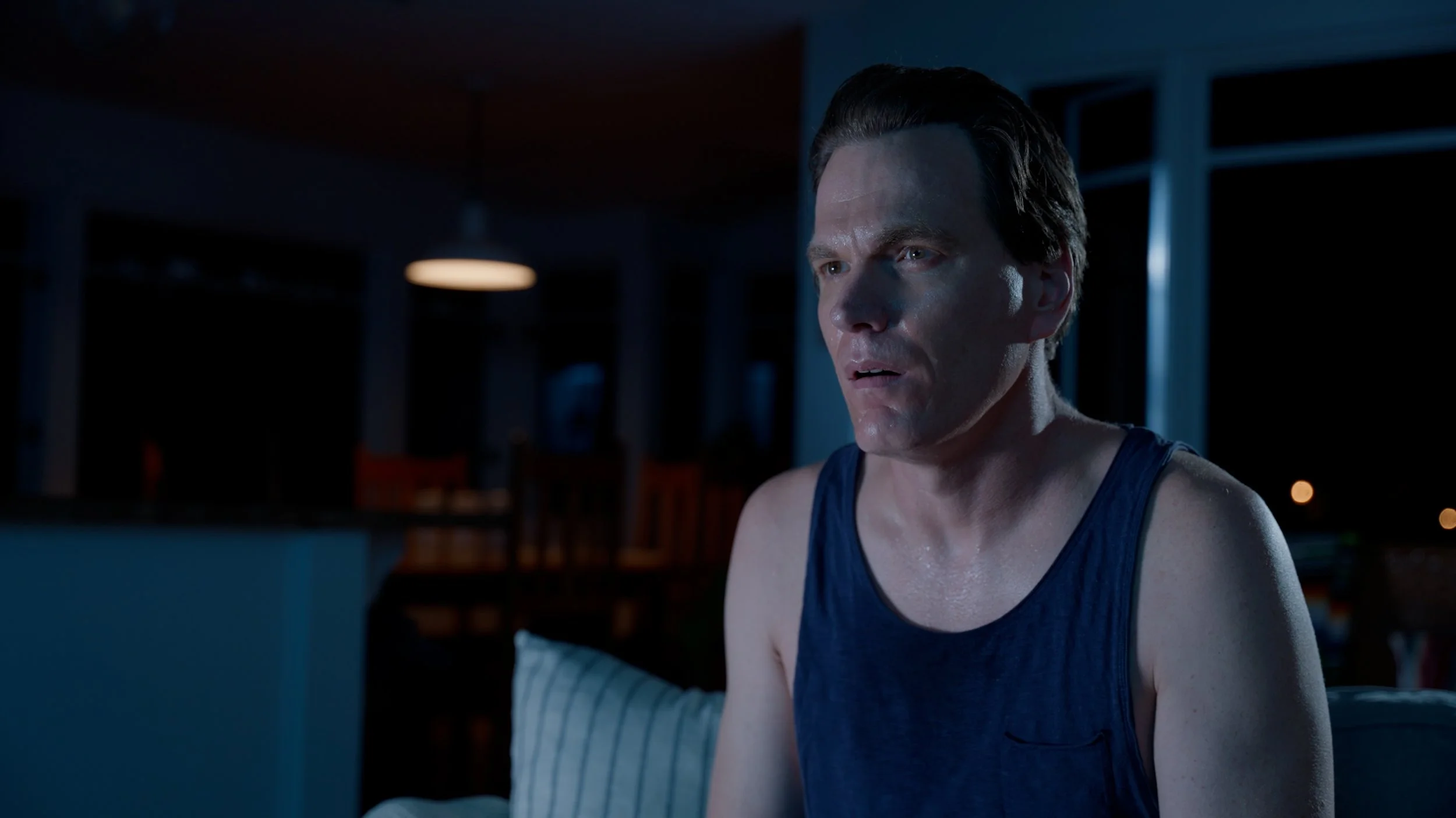 Man in dark room wearing blue tank top, looking intensely forward, with sweat on face.