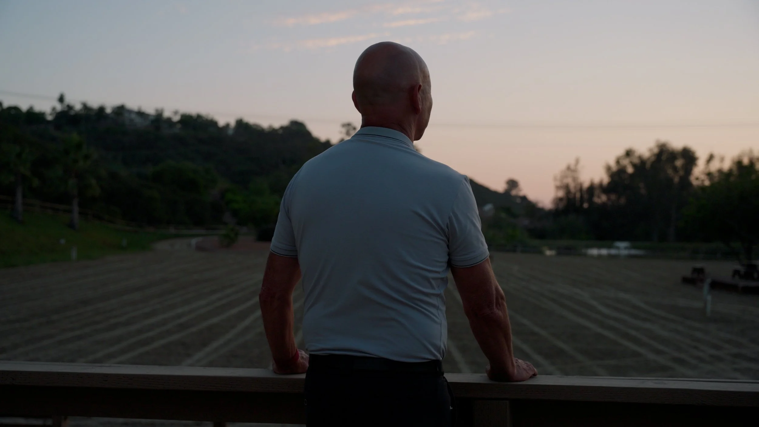 Person standing on a balcony overlooking a field and hills at sunset