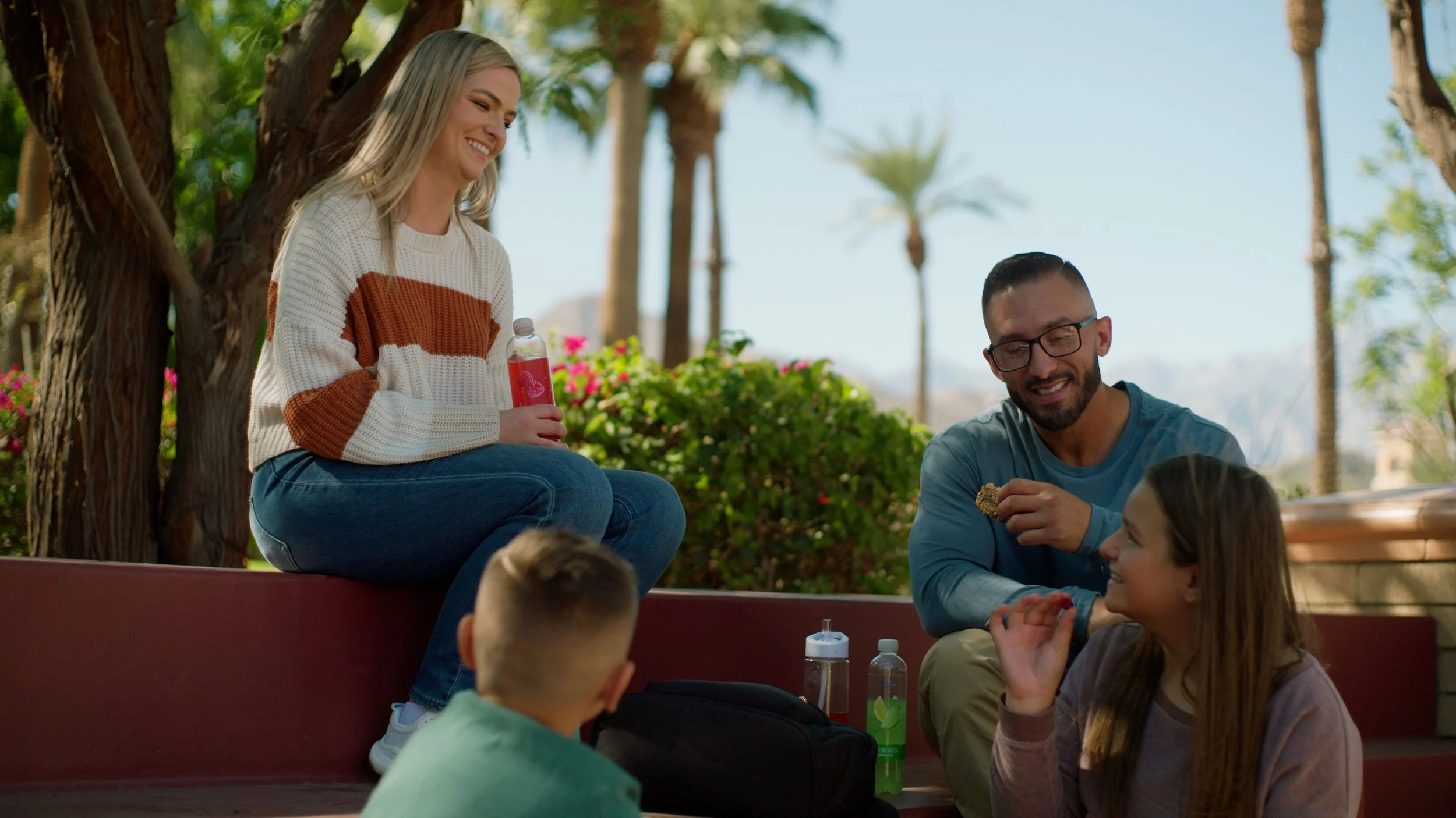 A group of people sitting outdoors near trees, smiling and talking, with beverages and snacks in hand.