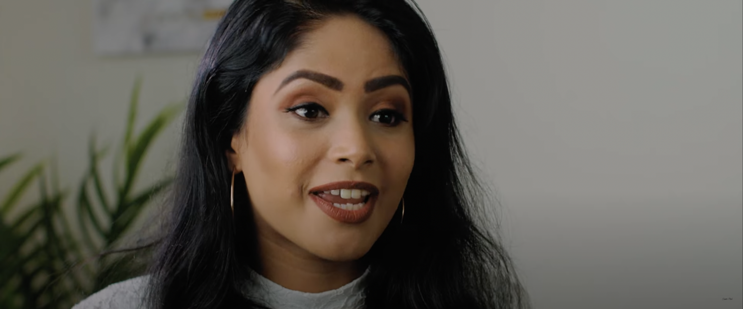 Woman speaking in an interview setting, indoor scene, with plants in the background.