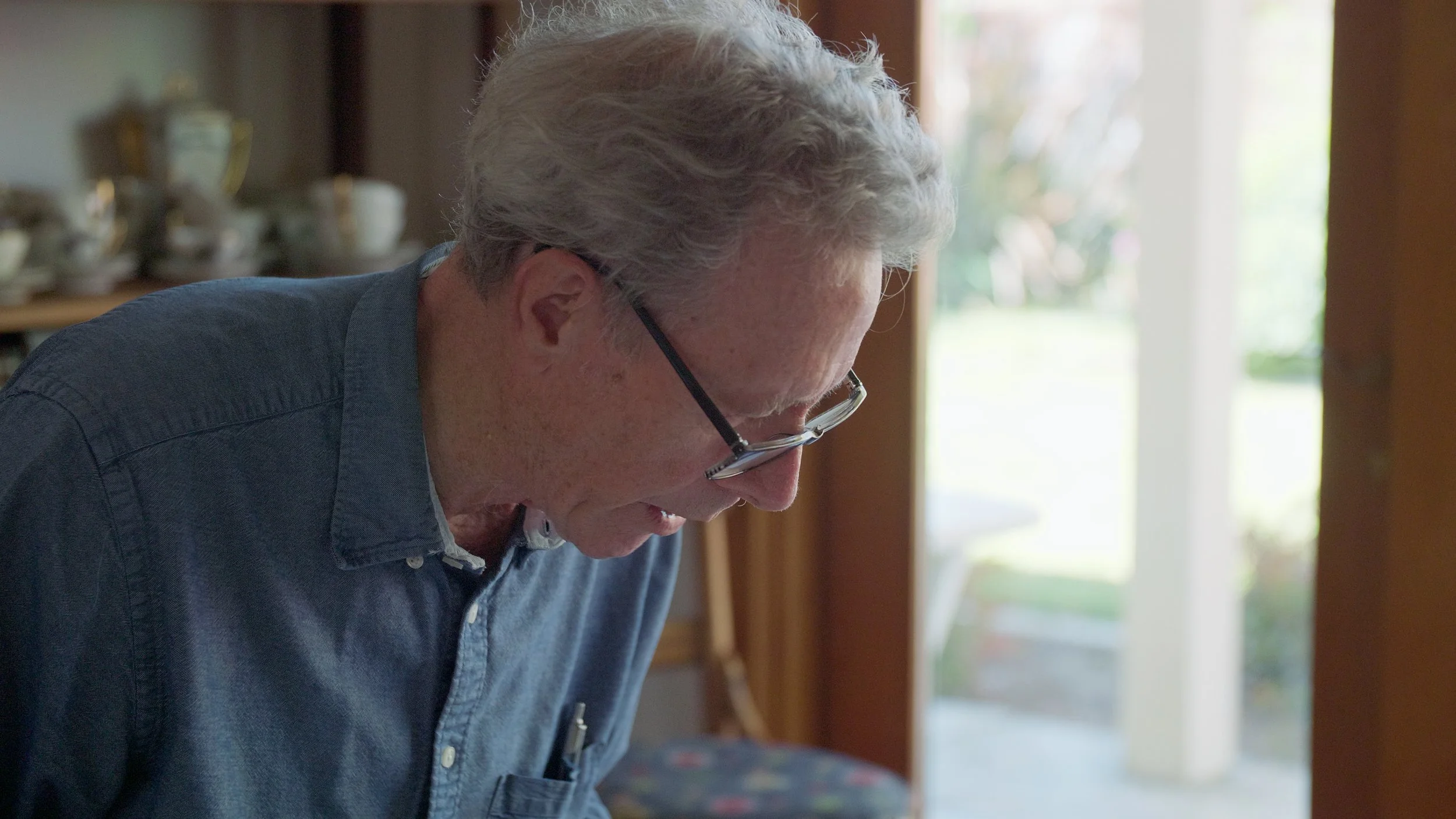 Elderly man in glasses and denim shirt looking down indoors