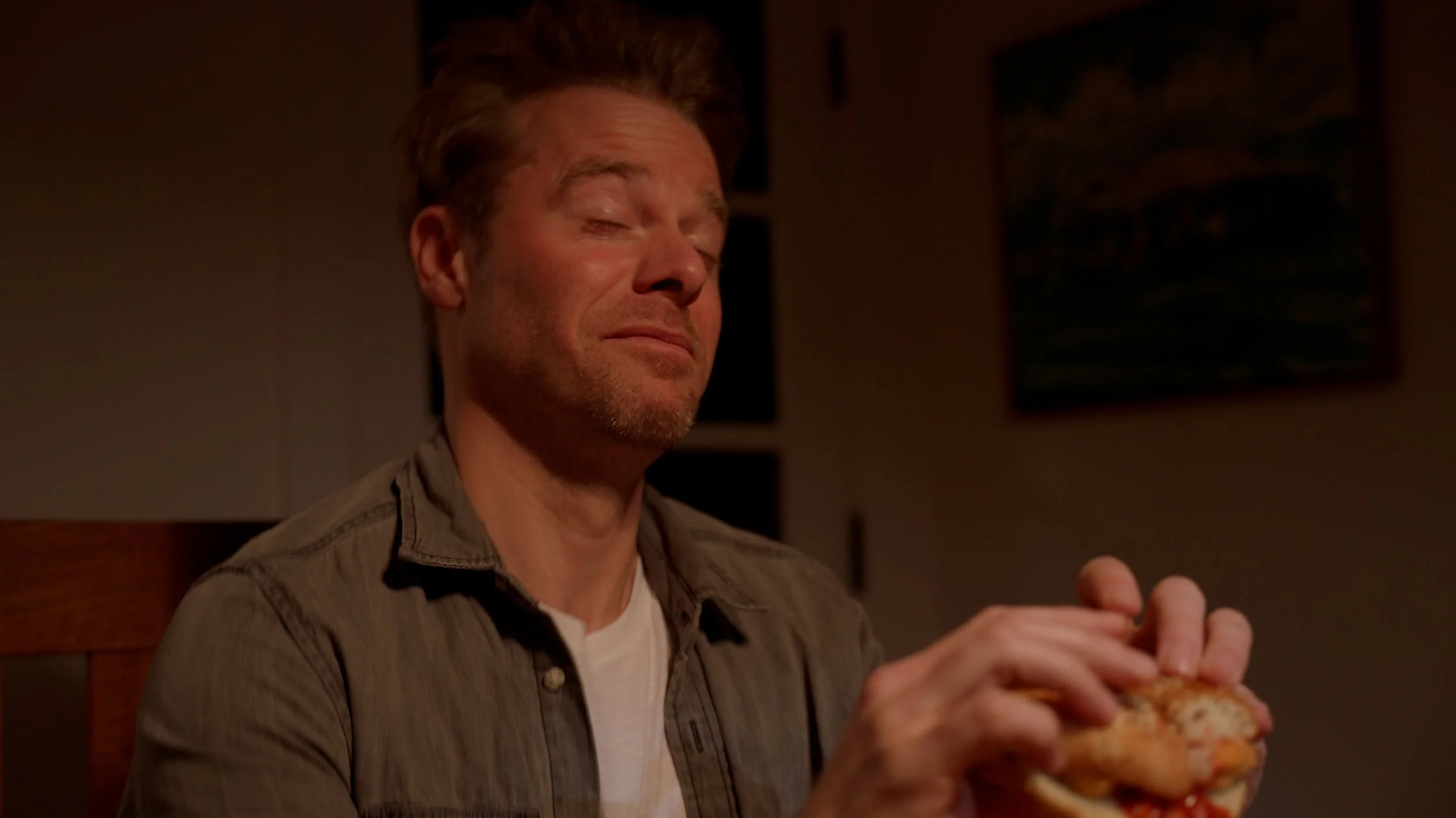 Person holding a sandwich with eyes closed, wearing a casual shirt.