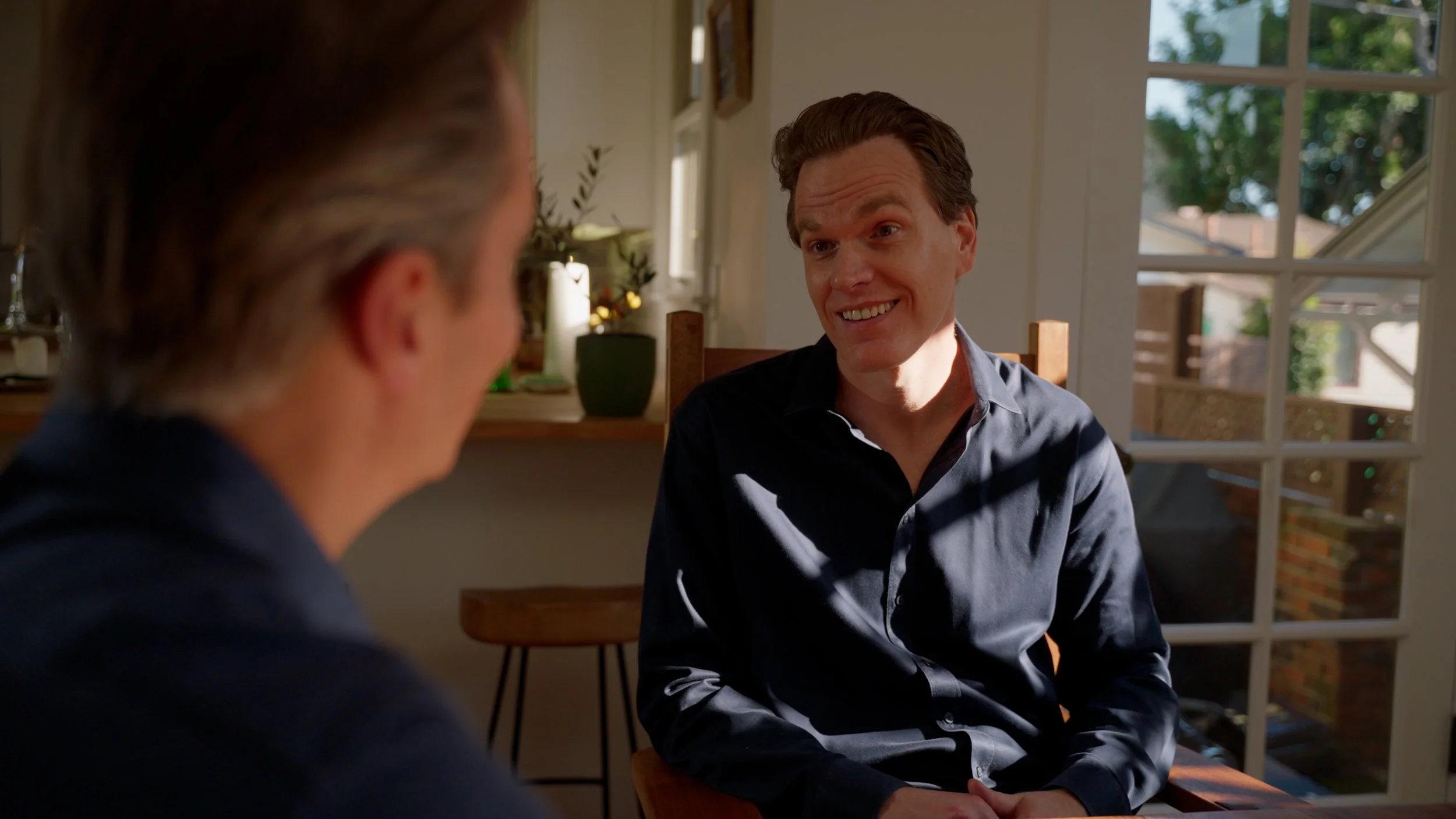 Two people sitting indoors having a conversation, one smiling, with natural light coming through the window.
