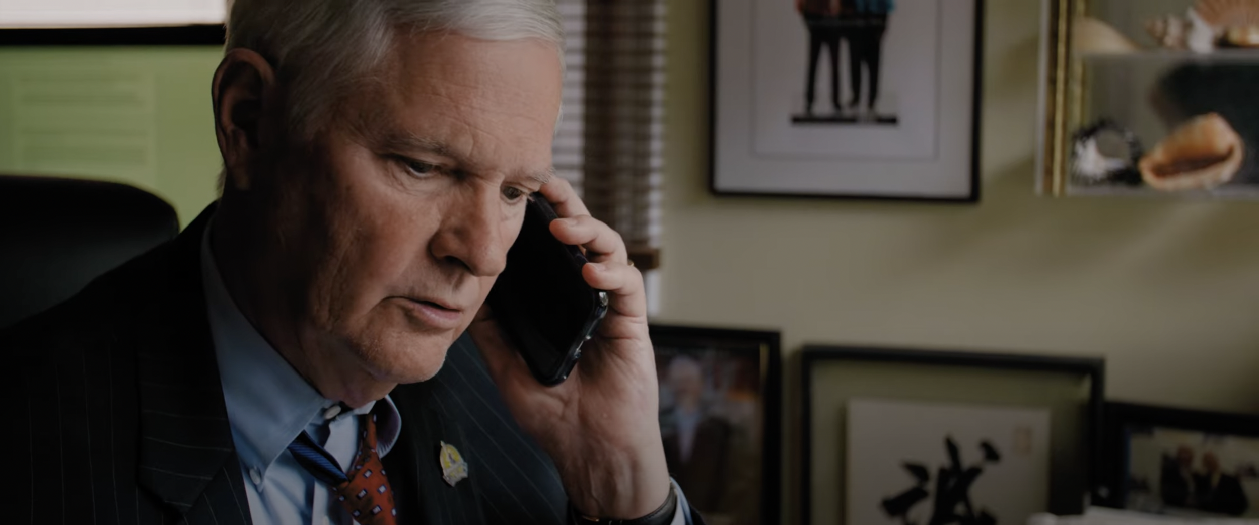 Man with gray hair on phone in office with framed photos and shelves with items