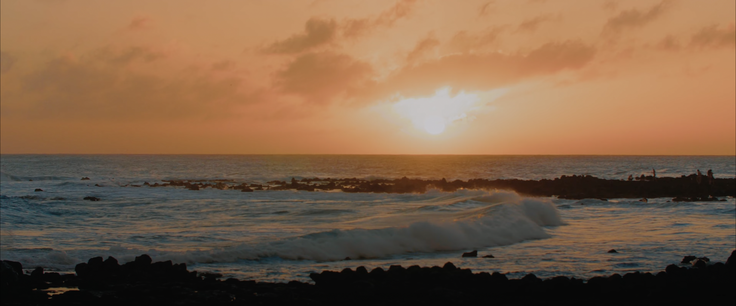 Sunset over ocean with waves and silhouette of rocky coastline.