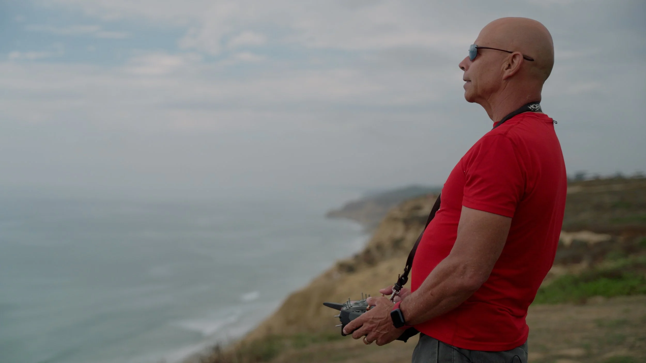 Person wearing a red shirt and sunglasses piloting a drone near a coastal cliff.