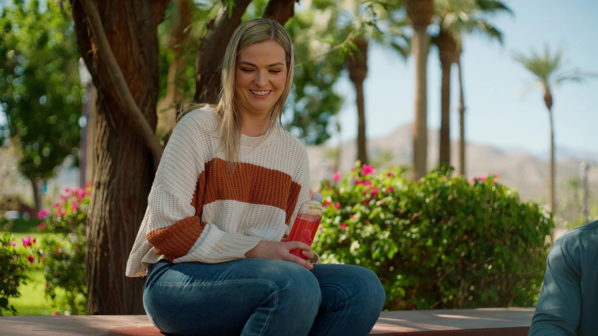 Smiling person sitting outside holding a pink drink, surrounded by trees and flowers.