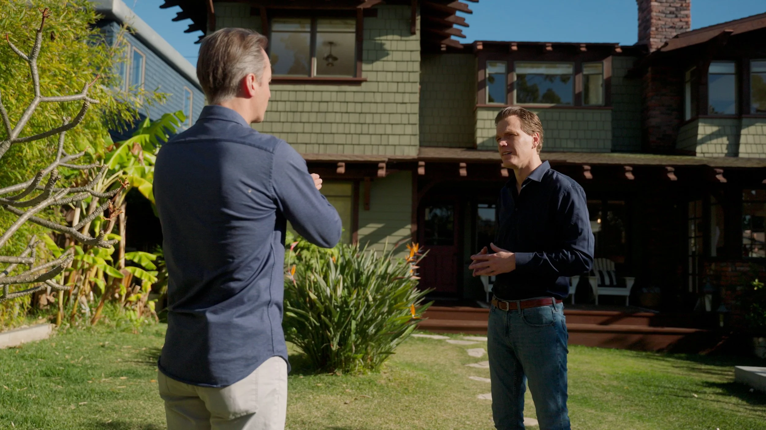 Two men talking in front of a traditional house with a shingled facade, lush garden, and clear blue sky.