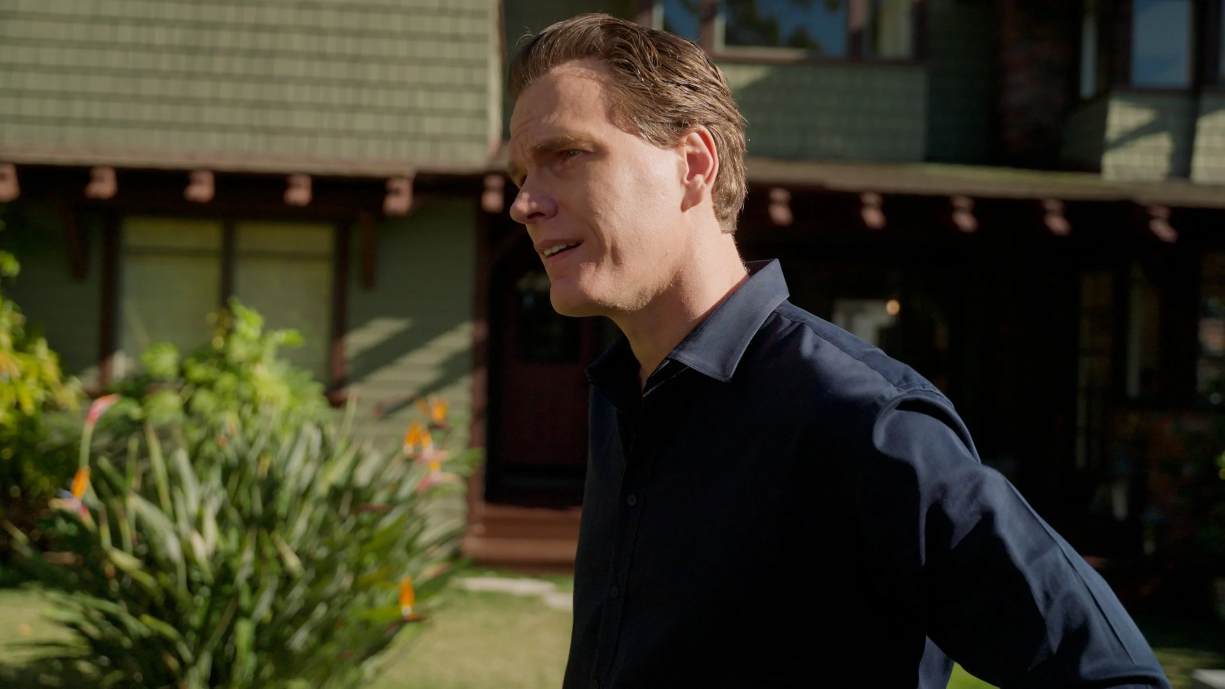 Man with short hair in a dark shirt standing outdoors in front of a house and plants.