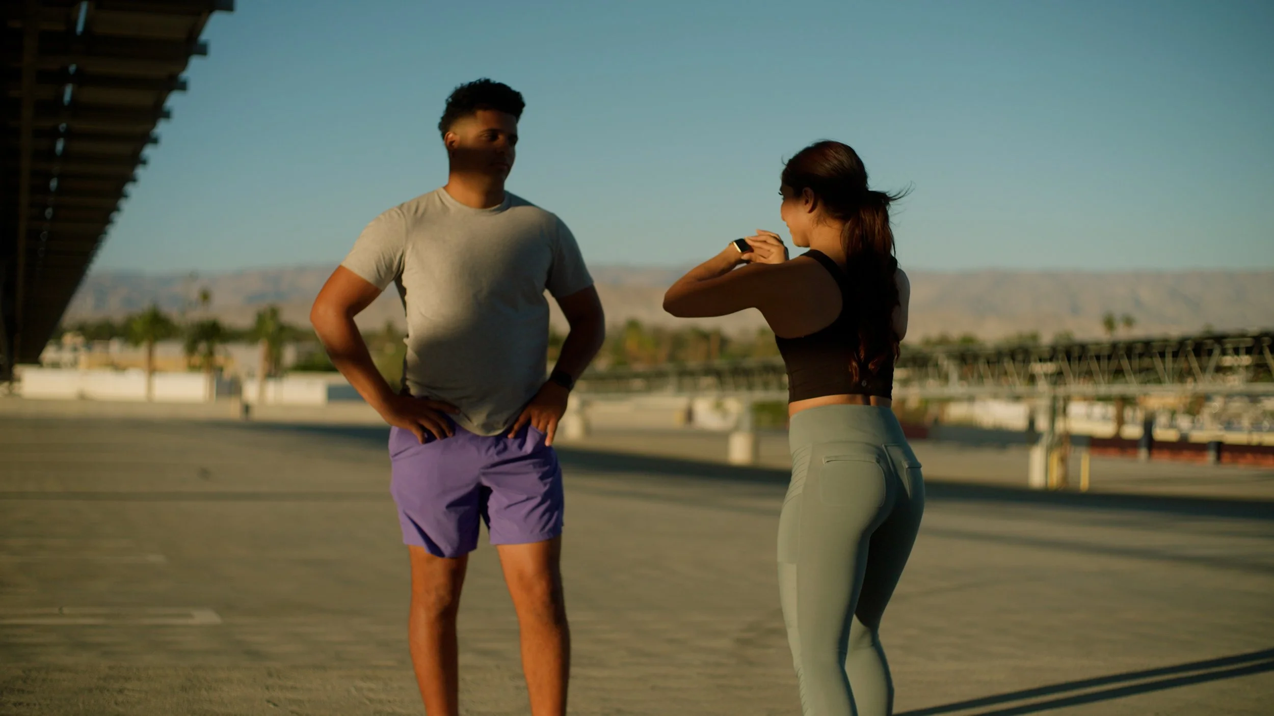 Two people exercising outdoors, one in gray shirt and purple shorts, the other in black tank top and light leggings.