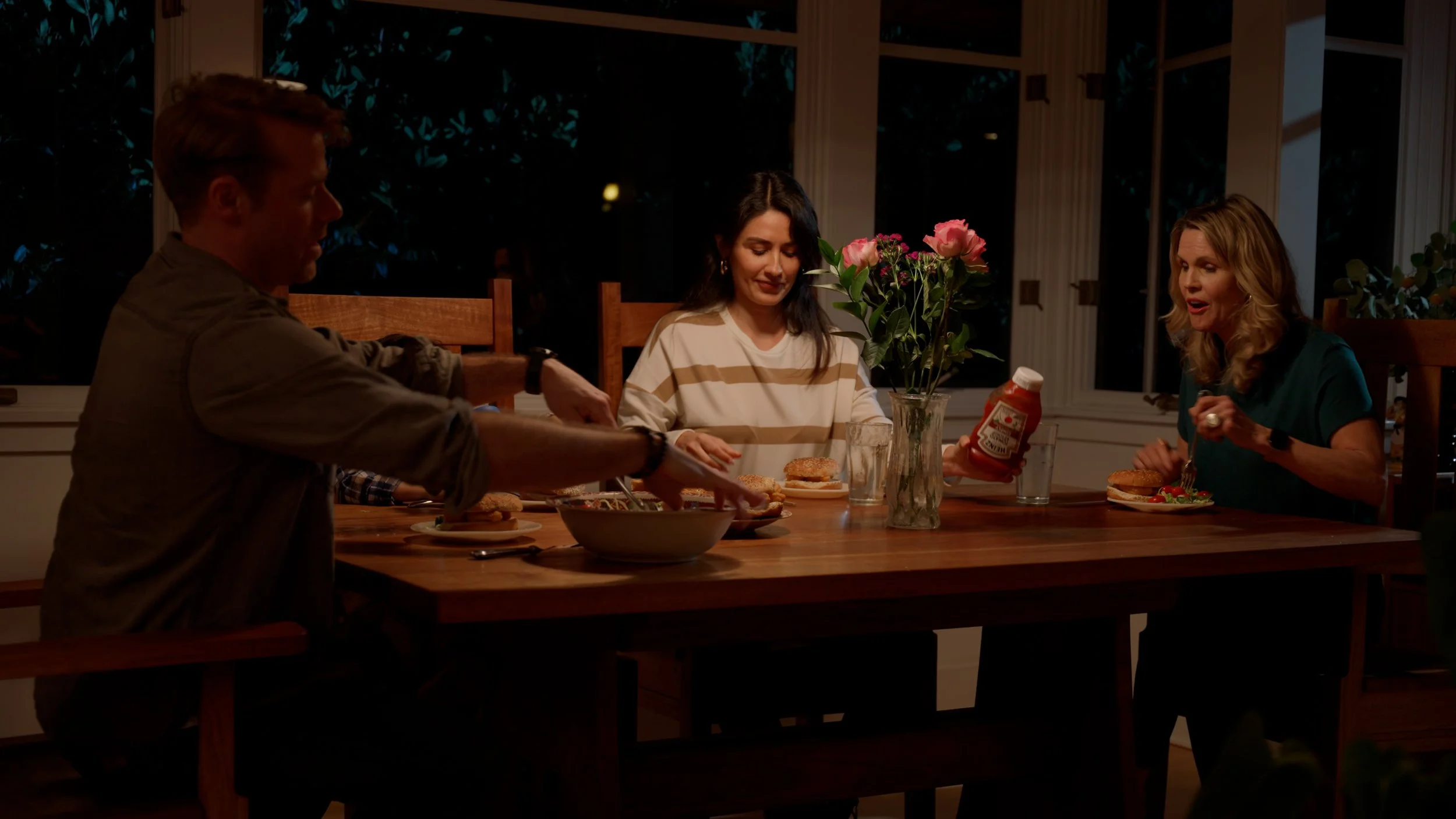 Family dinner table with four people, burgers, salad, and flowers in a vase.