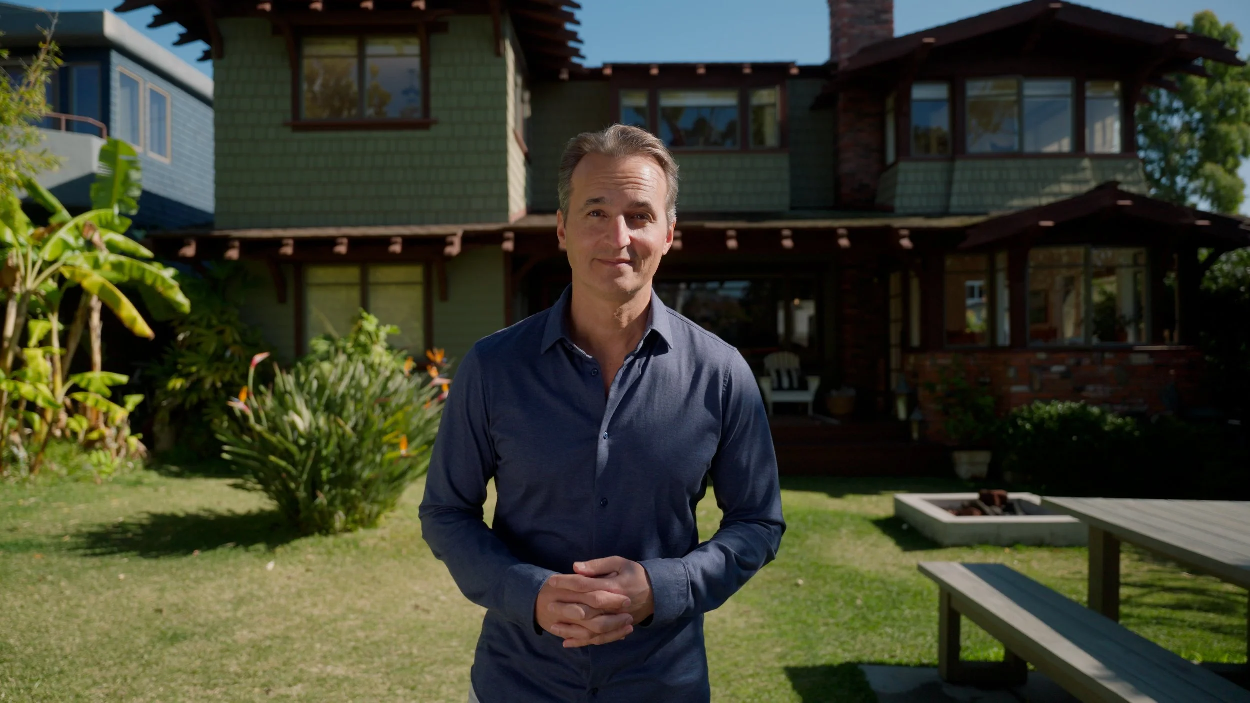 Man in blue shirt standing in front of a green two-story house with a garden.
