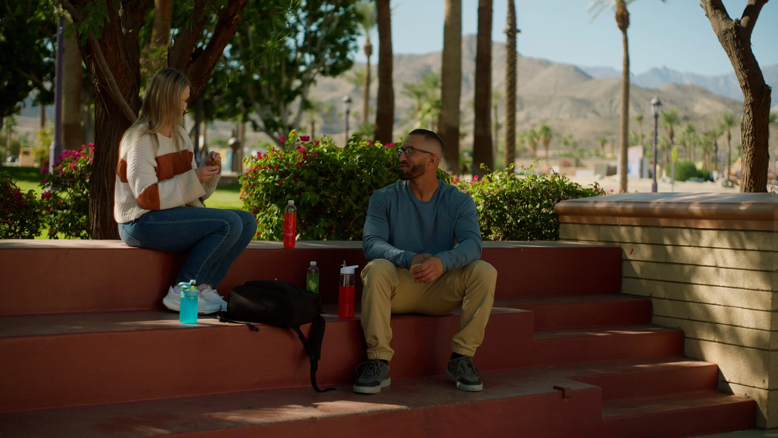Two people sitting on red steps outdoors with trees and mountains in the background, one holding a phone and the other looking over, surrounded by bags and water bottles.