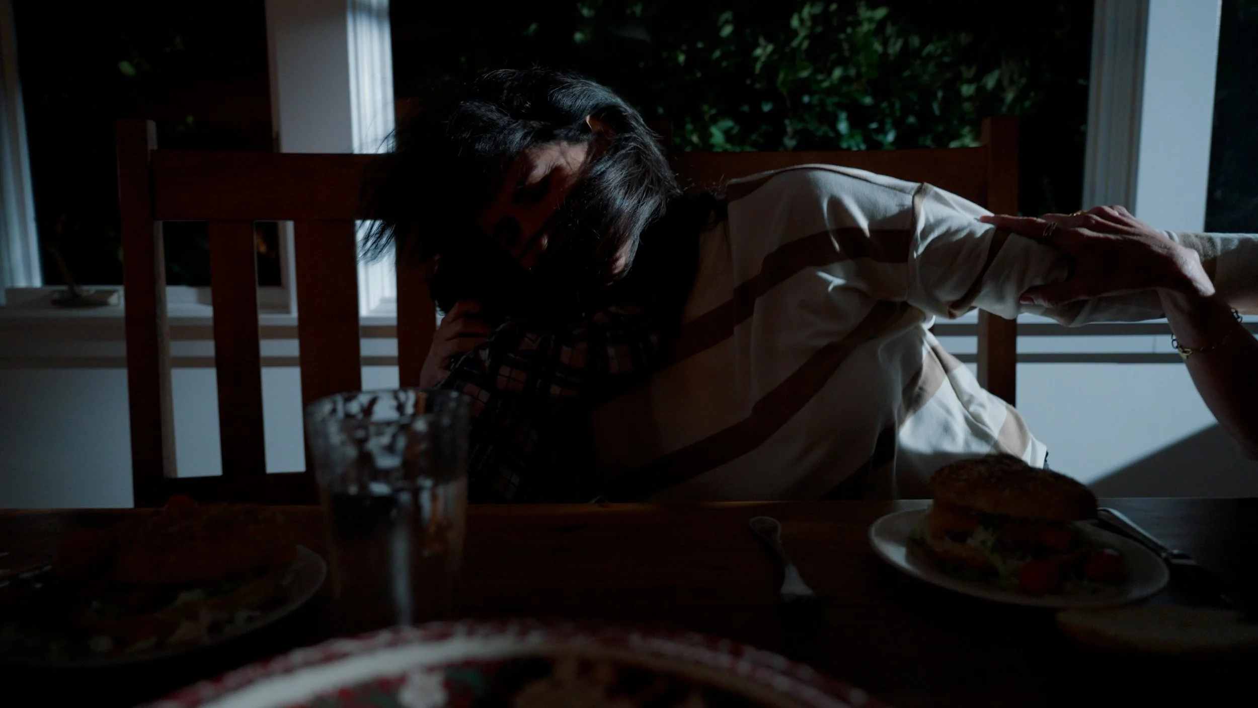 Dimly lit dining room scene with a person leaning on a chair, dark background, food plates, and an empty glass on a table.