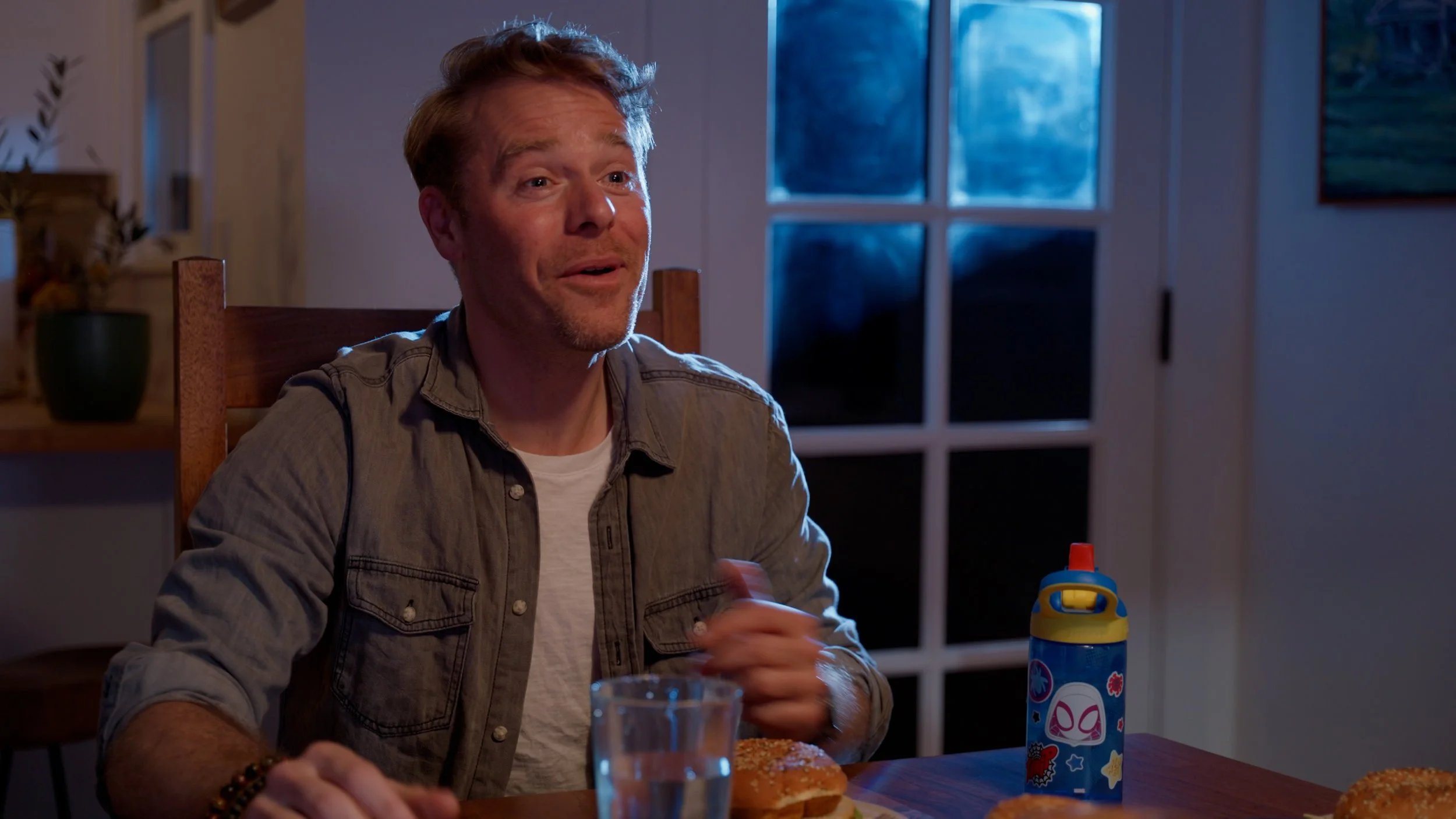 Man sitting at a table beside a window, smiling, with a water bottle decorated with superhero motifs.