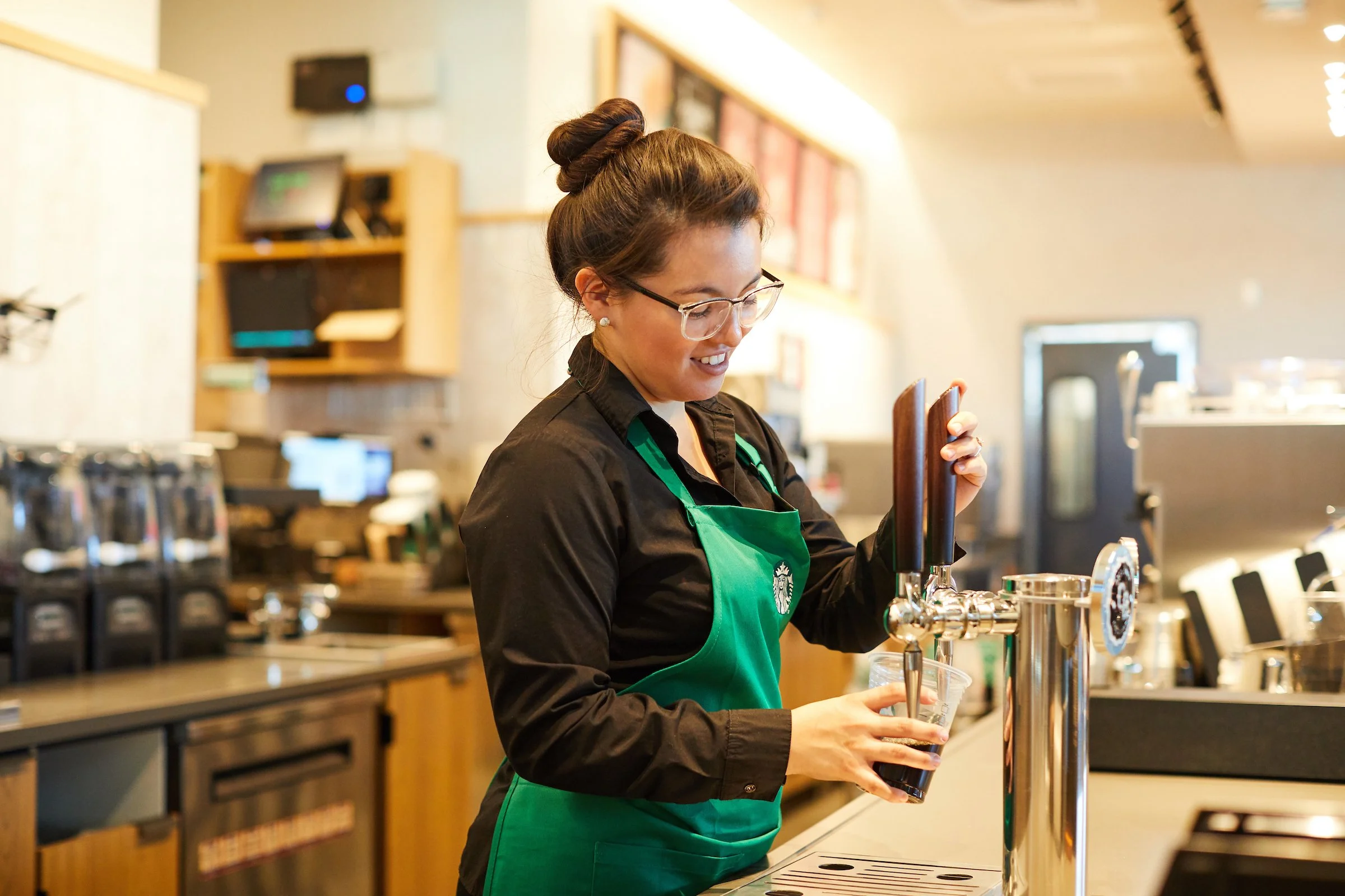 A woman in a black shirt and green apron pours a dark drink from a tap in a coffee shop.