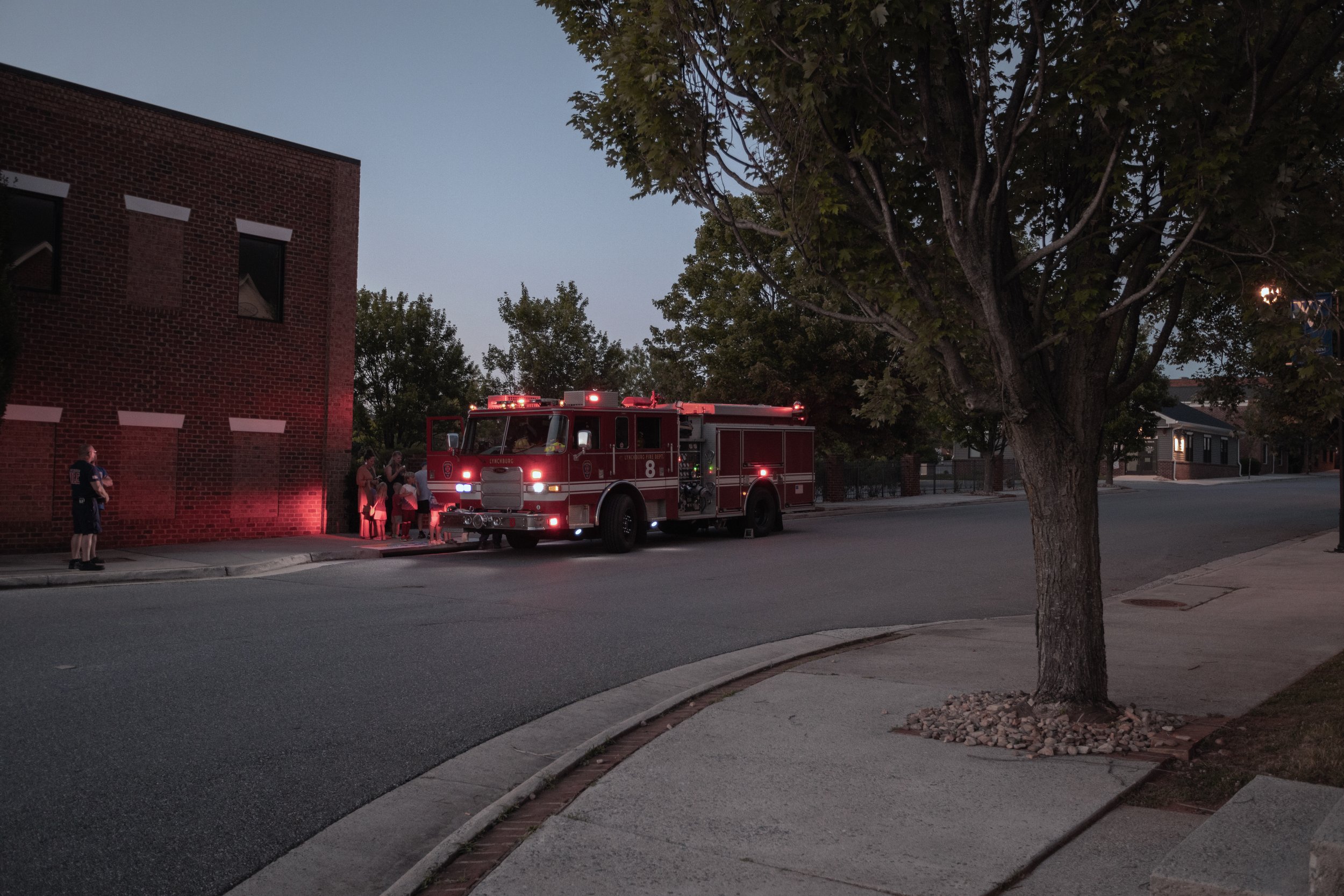 Fire truck parked on a quiet street at dusk with a group of people standing nearby, trees lining the street, and a brick building on the left side.