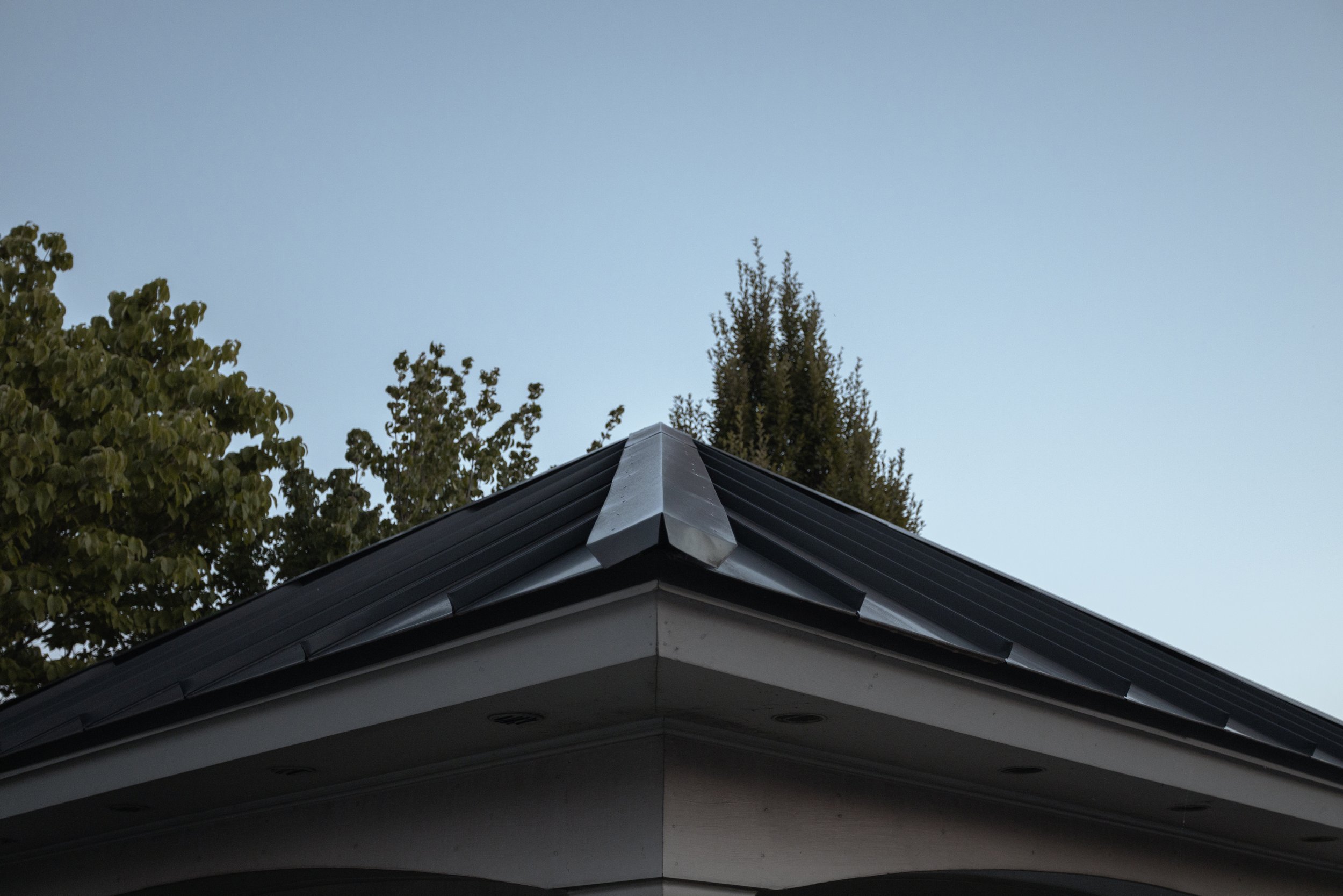 Close-up of a house roof with black shingles and metal flashing, trees in the background under a clear sky.