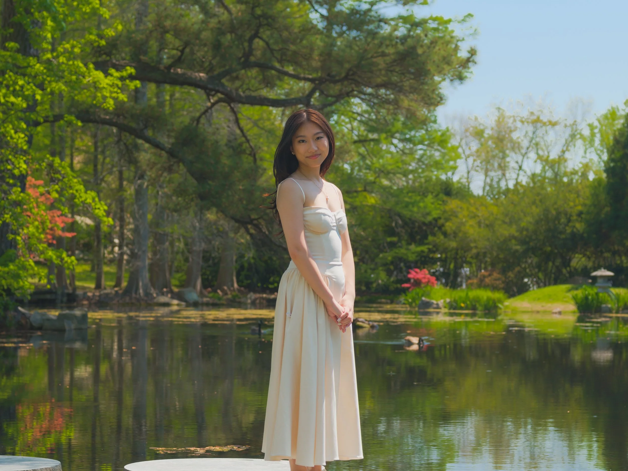 A young woman in a cream-colored dress standing by a pond in a lush garden with green trees and pink flowers, smiling at the camera.