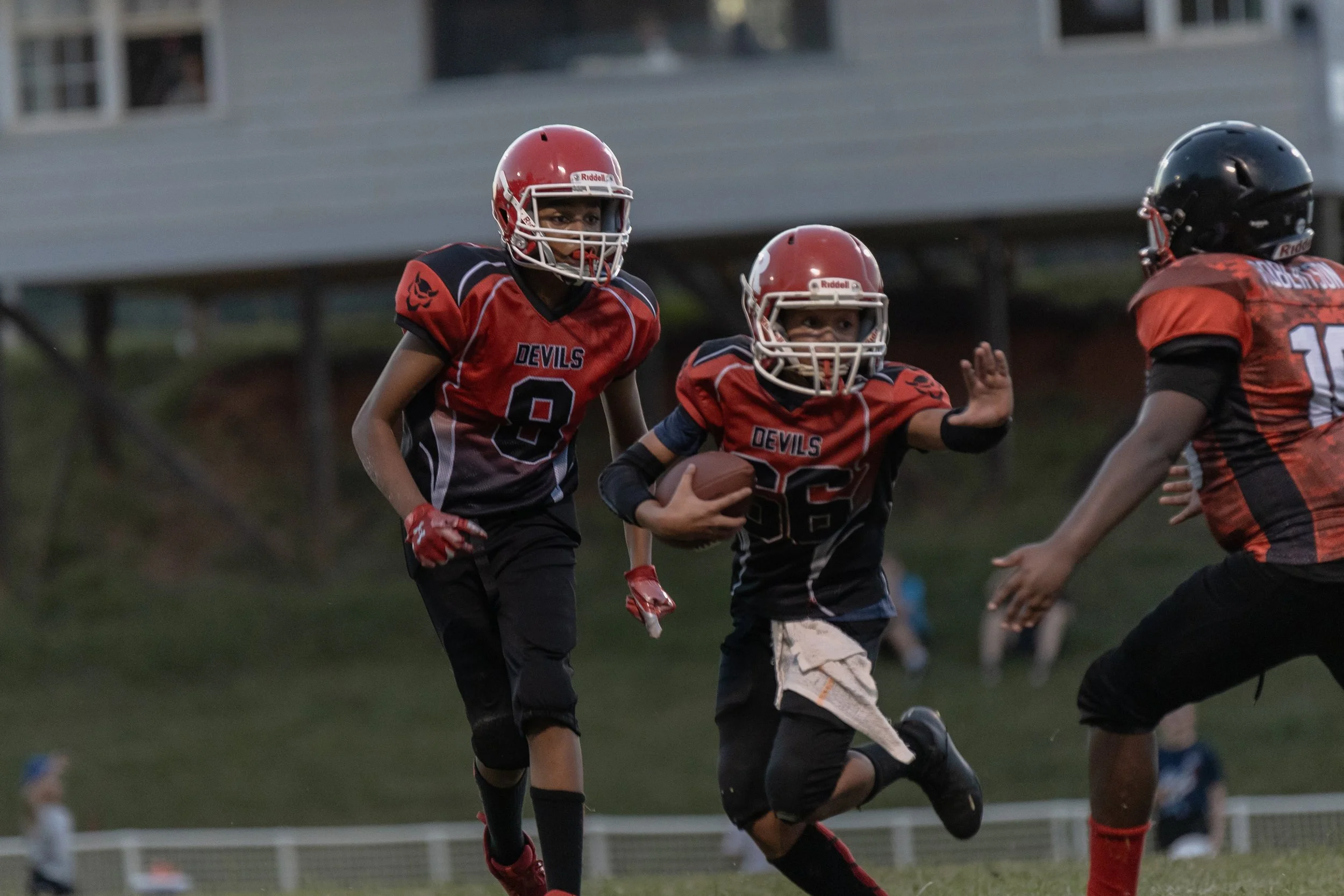 Young football players in red and black uniforms, with two wearing helmets, playing on a grassy field during a game. One player is running with the football while others pursue him.