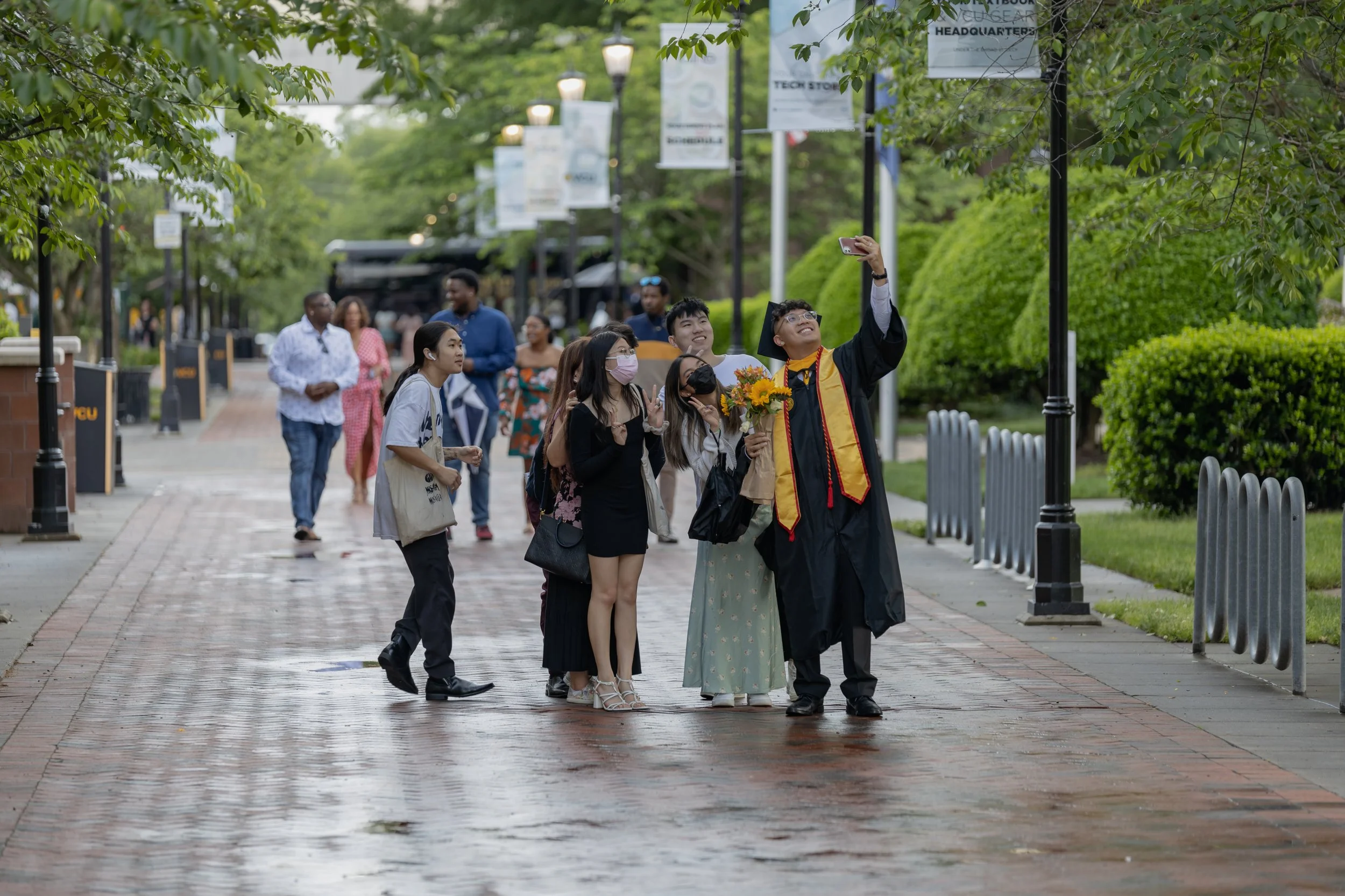 A group of people taking a photo of a graduate in cap and gown holding a bouquet during a graduation celebration on a brick-paved walkway with green trees and banners in the background.