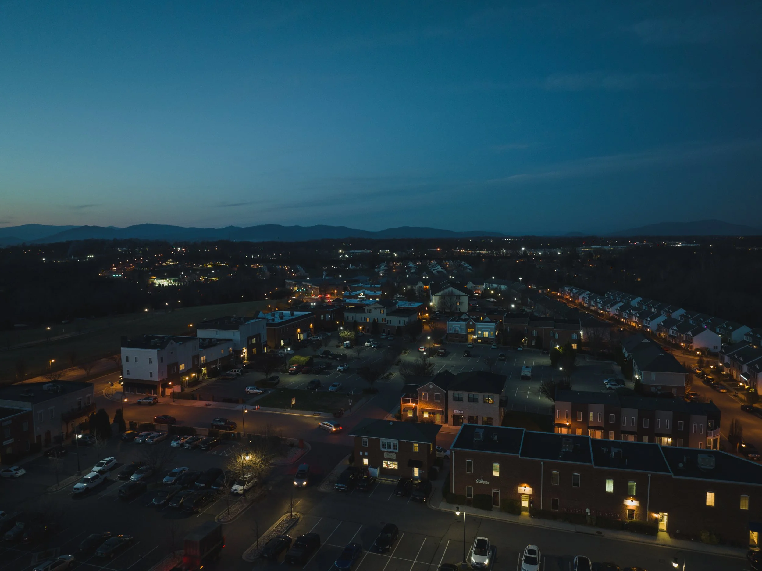 Dusk over a suburban neighborhood with illuminated houses and streets, with a mountain range in the distance.