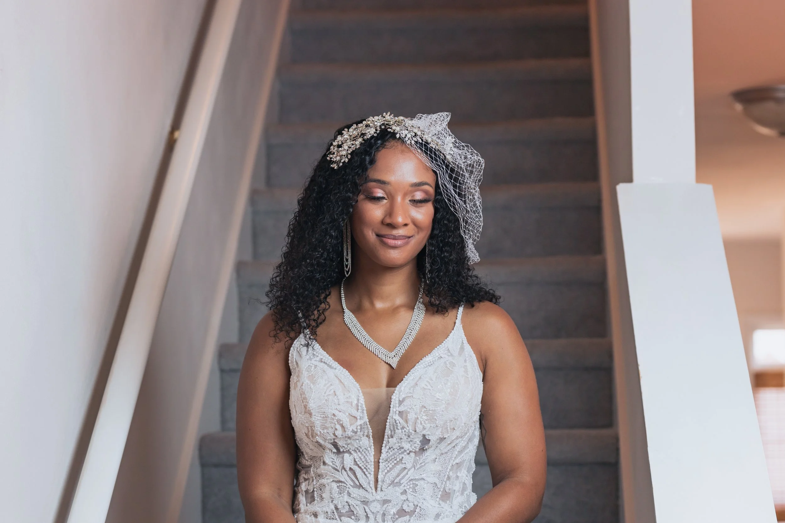 A woman dressed in a wedding gown with lace details, wearing a pearl necklace, earrings, and a decorative headpiece with a birdcage veil, standing on a staircase with a serene expression.