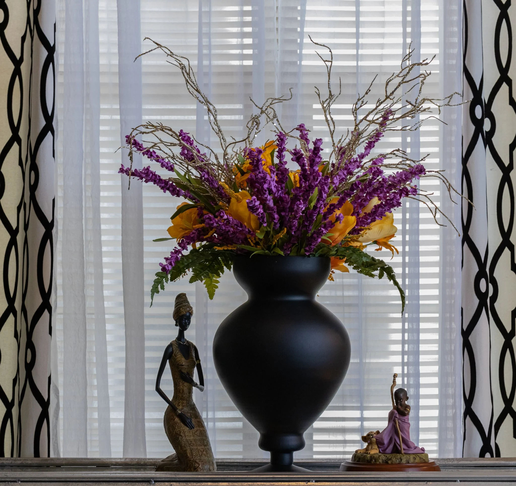 A large black vase with purple and yellow flowers, including tall purple flowers and yellow calla lilies, on a table in front of a window with sheer curtains and black-and-white patterned drapes. Two decorative figurines, one of a woman and the other