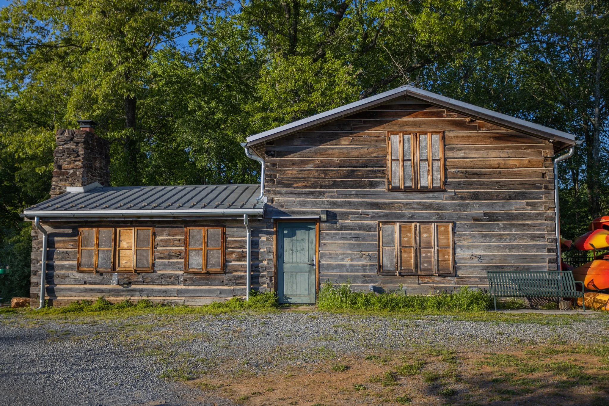 Old wooden house with boarded-up windows, a chimney, a metal roof, surrounded by green trees.