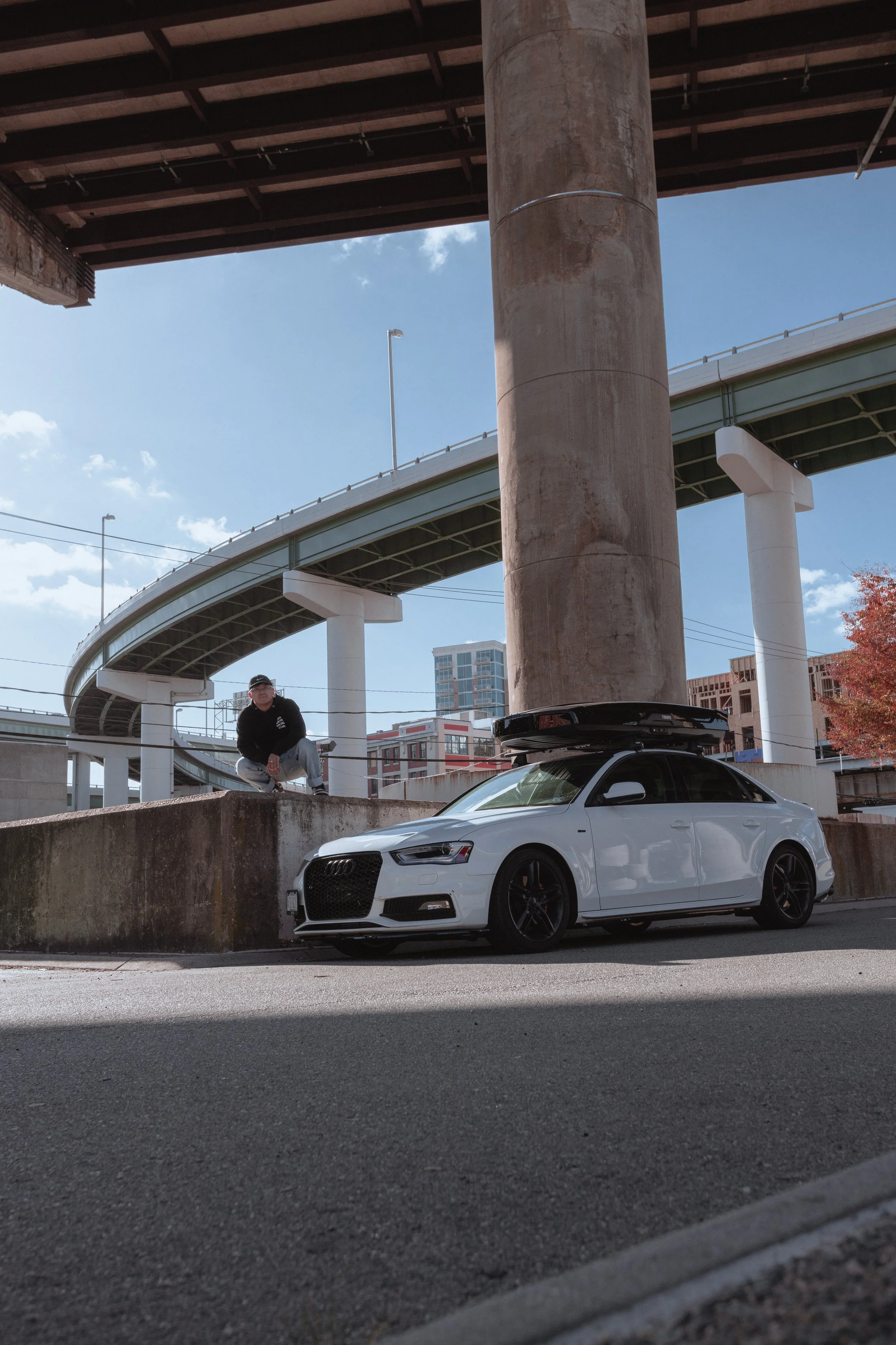 A man crouching on a low concrete wall next to a white Audi sedan with a roof cargo box, under an overpass in an urban setting.