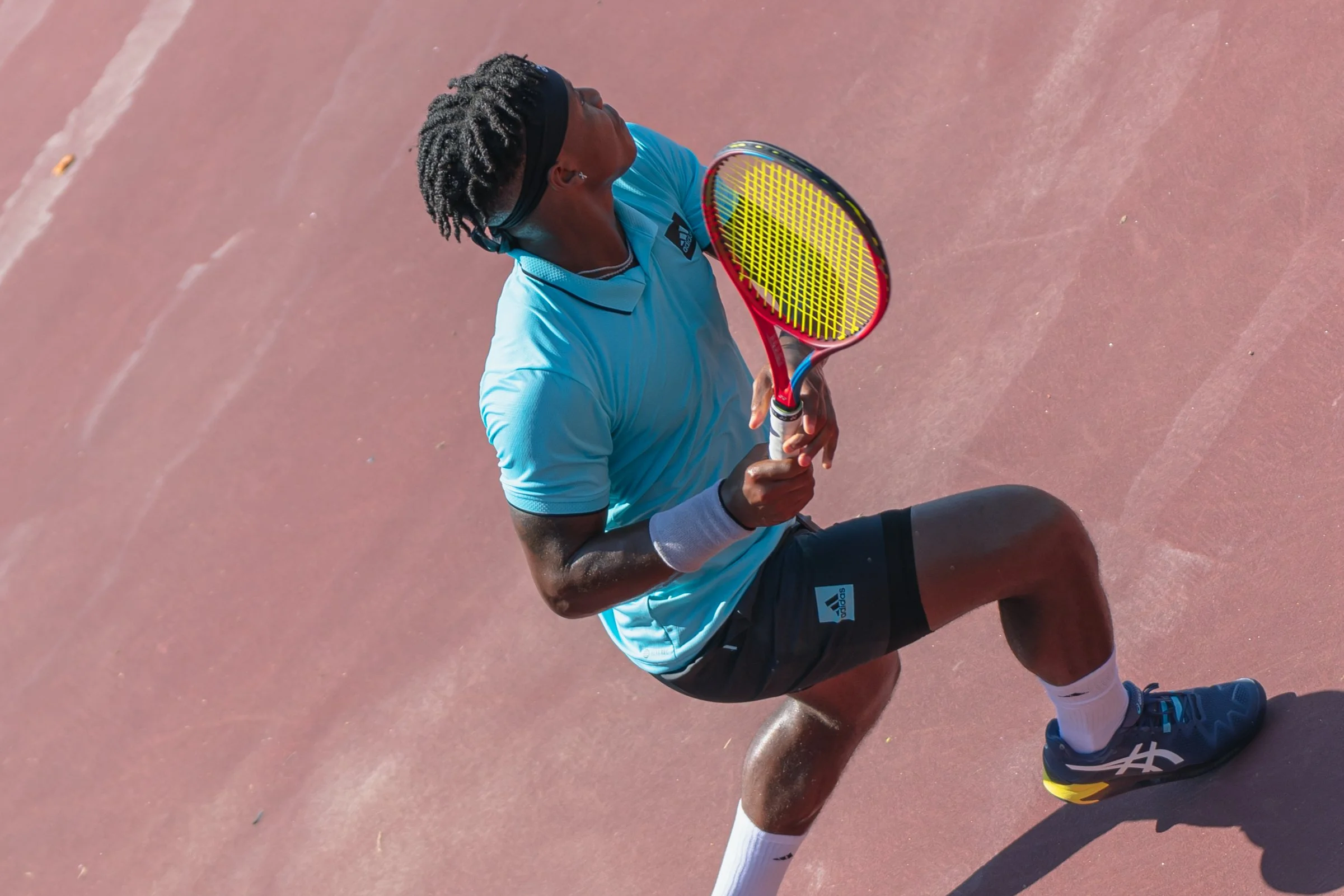 Male tennis player in blue shirt, black shorts, white socks, and blue shoes, with a black headband, holding a tennis racket on a red clay court.