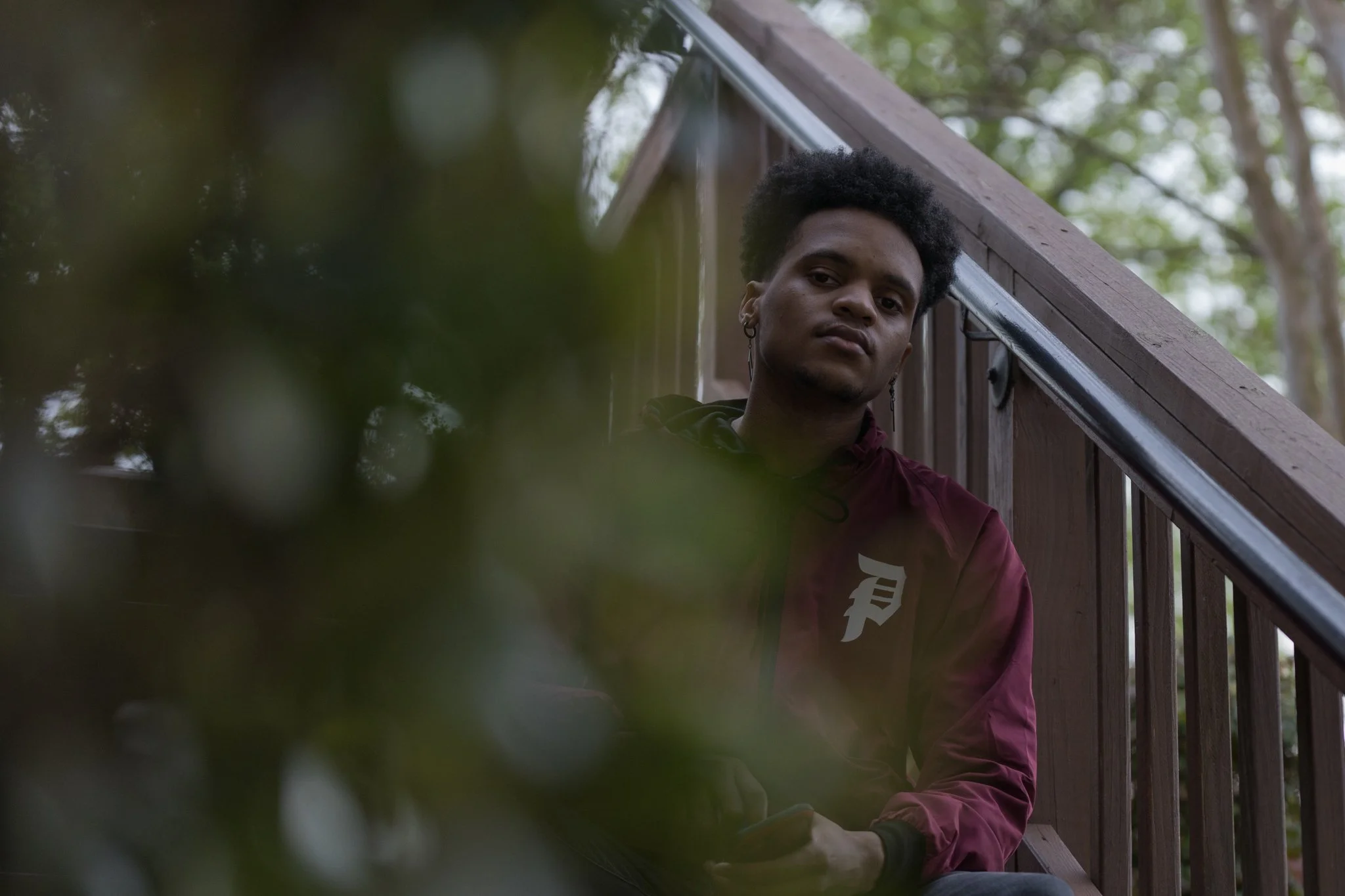 Young man with dark curly hair and earrings sitting on a staircase outside, partially obscured by blurred leaves in the foreground.