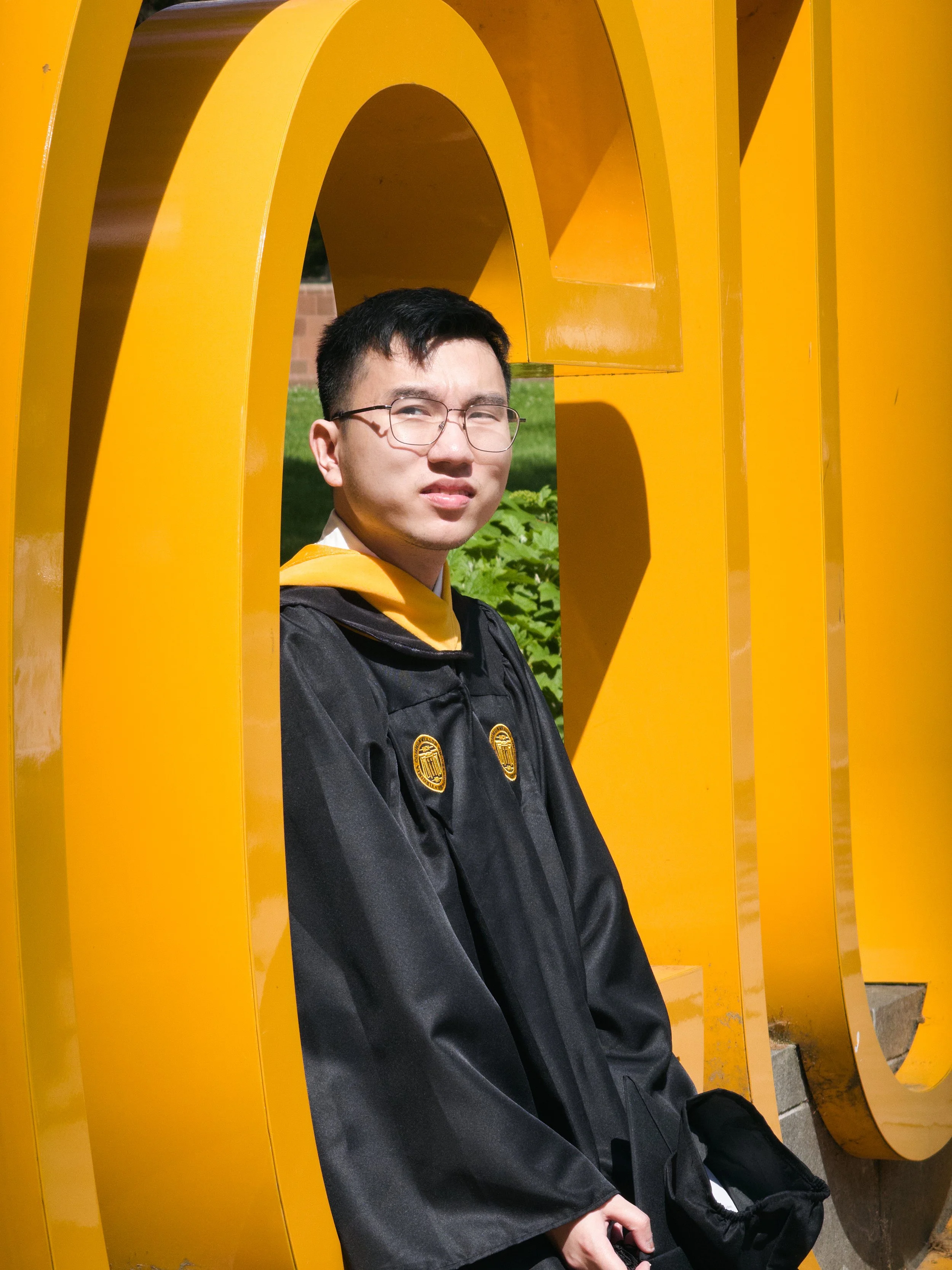 A young man wearing glasses and a black graduation gown stands in front of a large yellow sculpture, squinting his eyes in the sunlight.