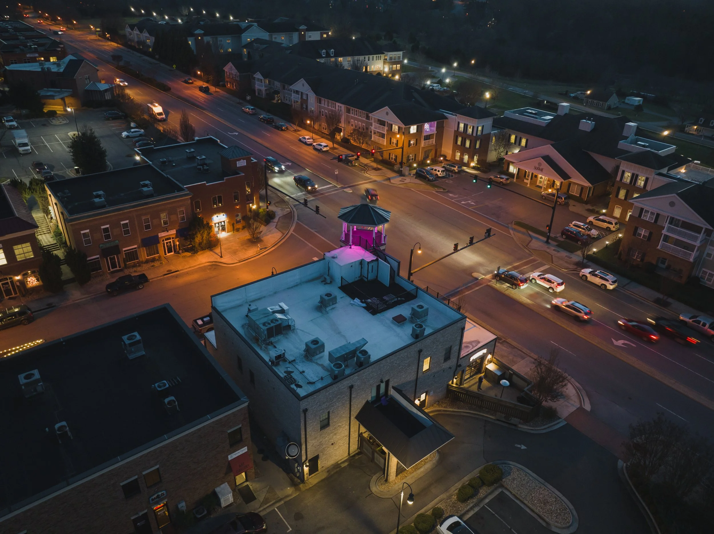 Nighttime aerial view of a downtown area with a small park featuring a gazebo with pink lighting, surrounded by parking lots and buildings. Traffic is moving on the main streets, and the area is illuminated by streetlights.